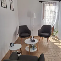 Jasny gabinet z trzema szarymi fotelami, małymi okrągłymi stolikami, lampą stojącą, zasłoniętym oknem i dekoracjami na ścianach, tworzący przytulną atmosferę.