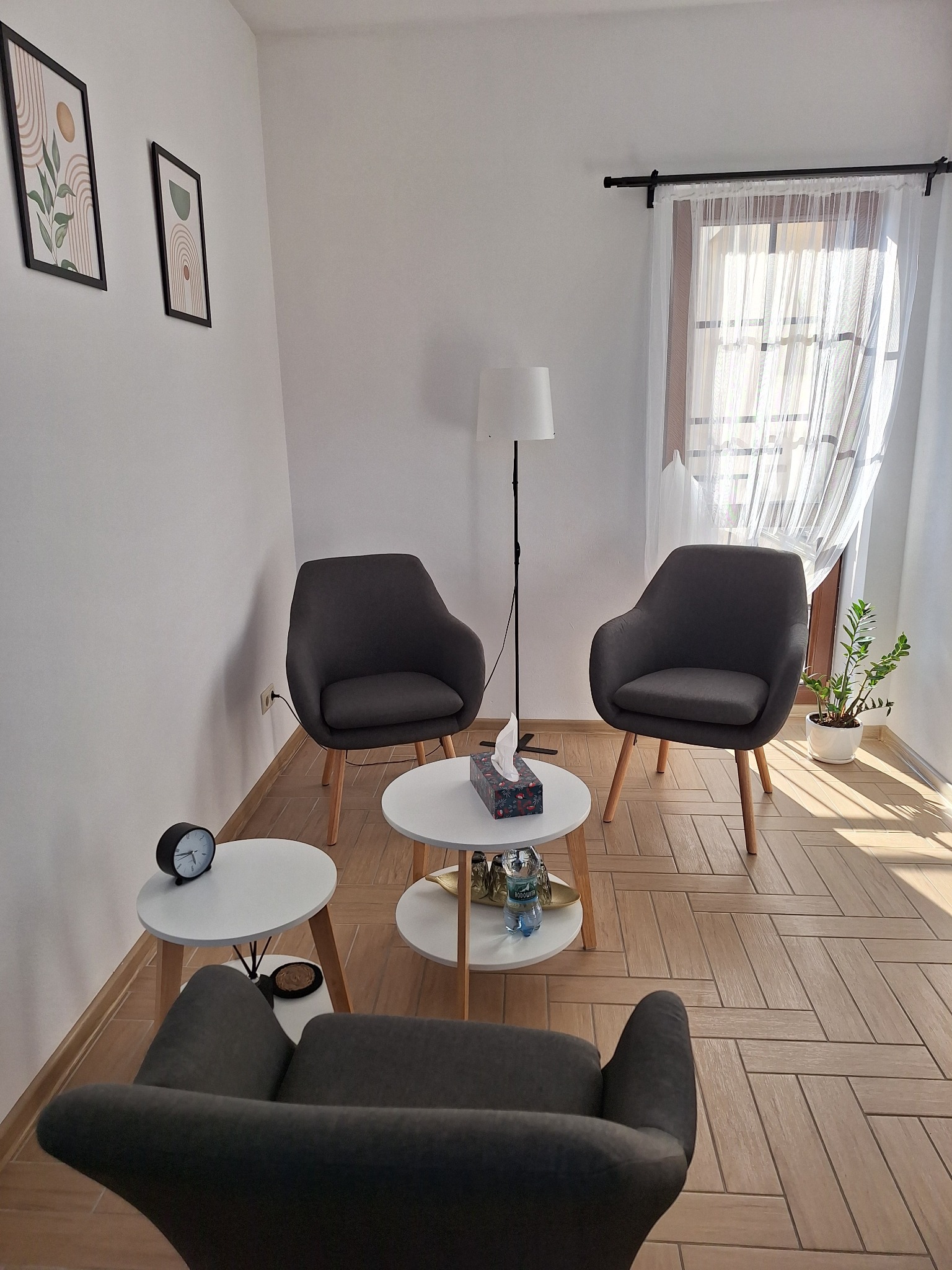 Jasny gabinet z trzema szarymi fotelami, małymi okrągłymi stolikami, lampą stojącą, zasłoniętym oknem i dekoracjami na ścianach, tworzący przytulną atmosferę.