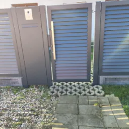 Nowoczesna, szara brama z poziomymi lamelami i furtka z wideodomofonem, zamontowana na posesji z kamiennym podłożem i betonowymi płytami.