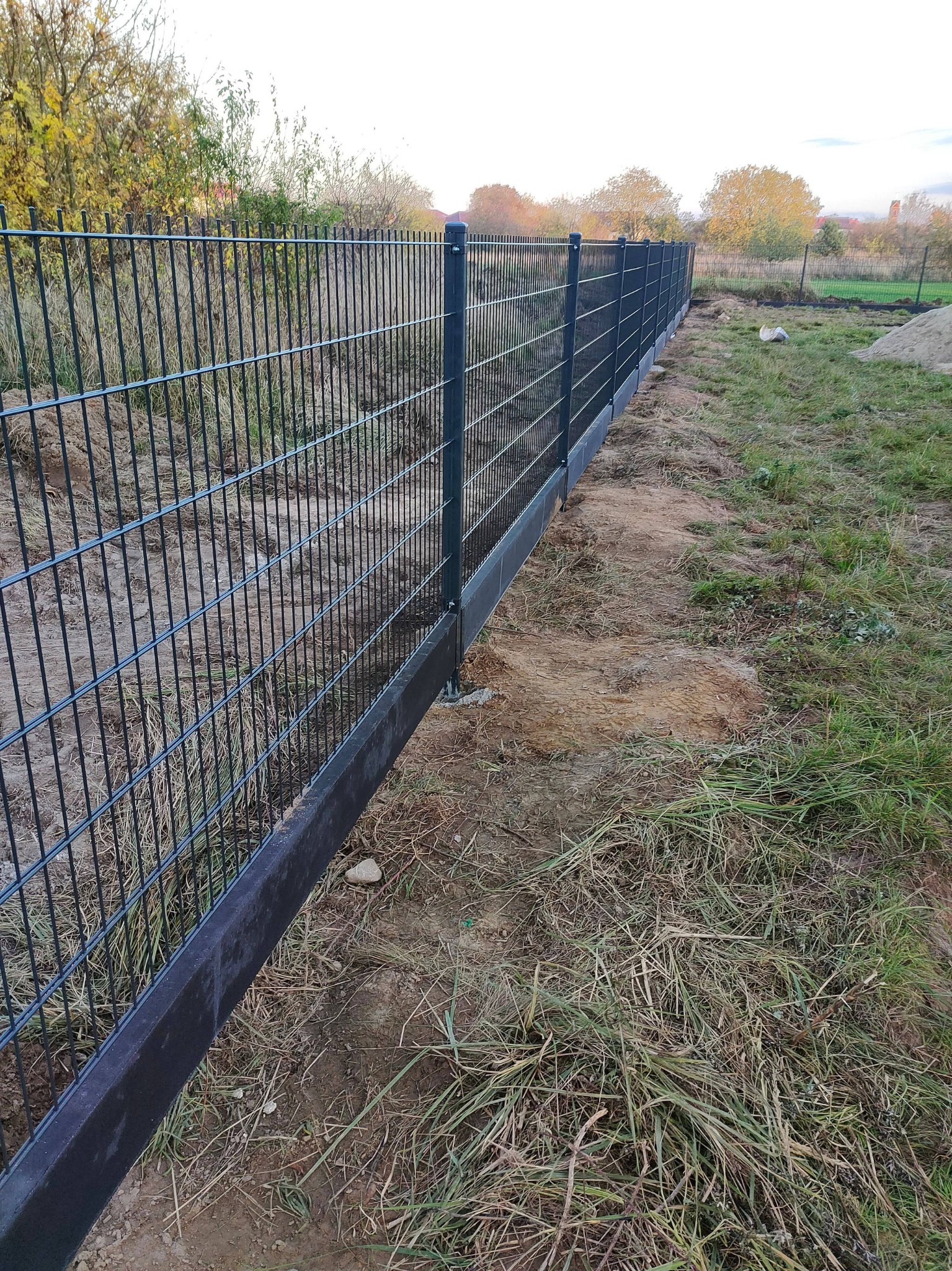 Nowoczesne, ciemne ogrodzenie z paneli stalowych z podmurówką betonową, widok z boku na tle jesiennego krajobrazu.