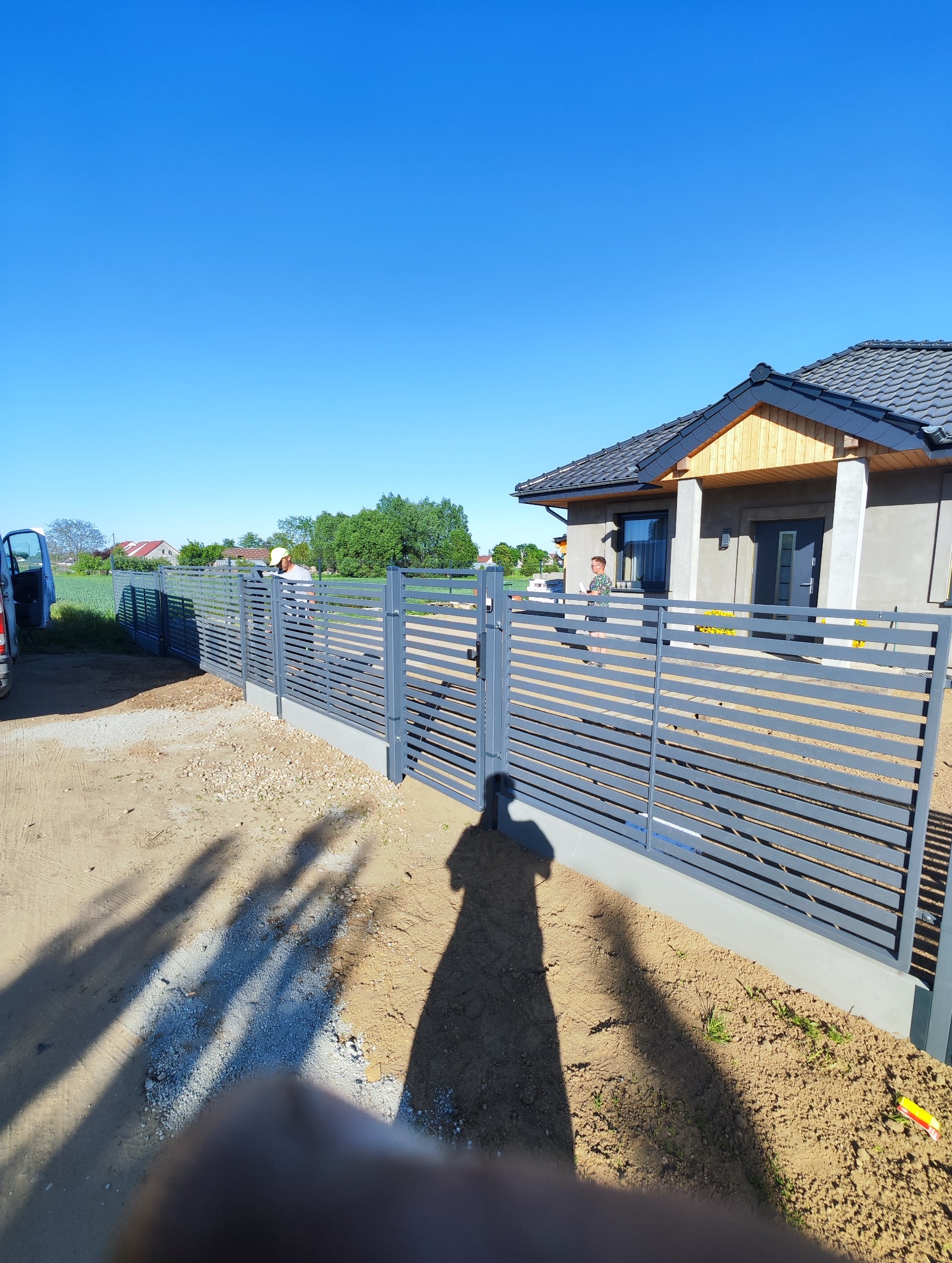 Nowoczesne, poziome, szare przęsła ogrodzeniowe z bramą i furtką, montowane przez dwóch pracowników w białych czapkach, na tle domu z szarą elewacją i niebieskim niebem.