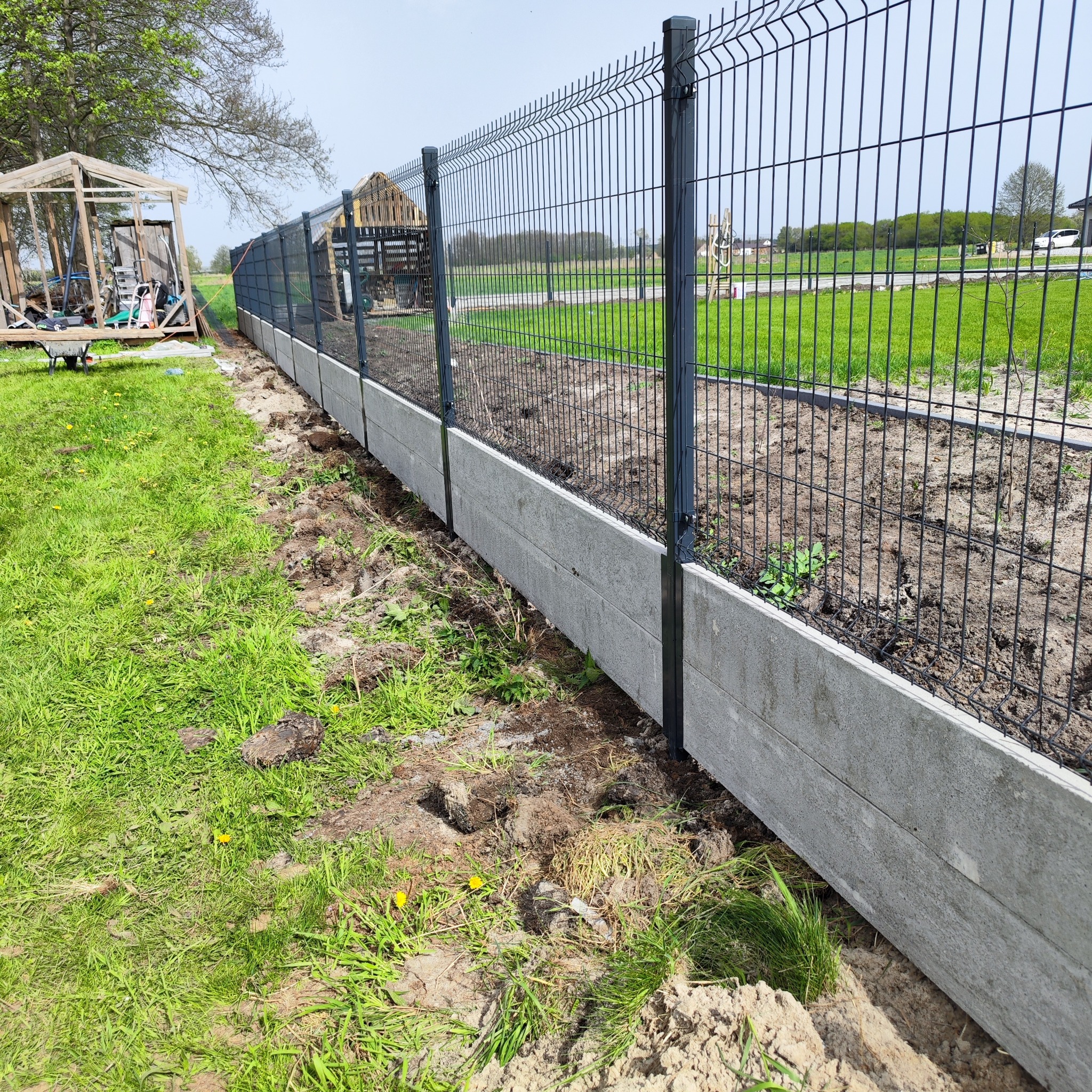 Nowo postawione czarne ogrodzenie panelowe z betonową podmurówką na nierównym terenie z widocznymi śladami prac ziemnych i fragmentem drewnianej konstrukcji w tle.