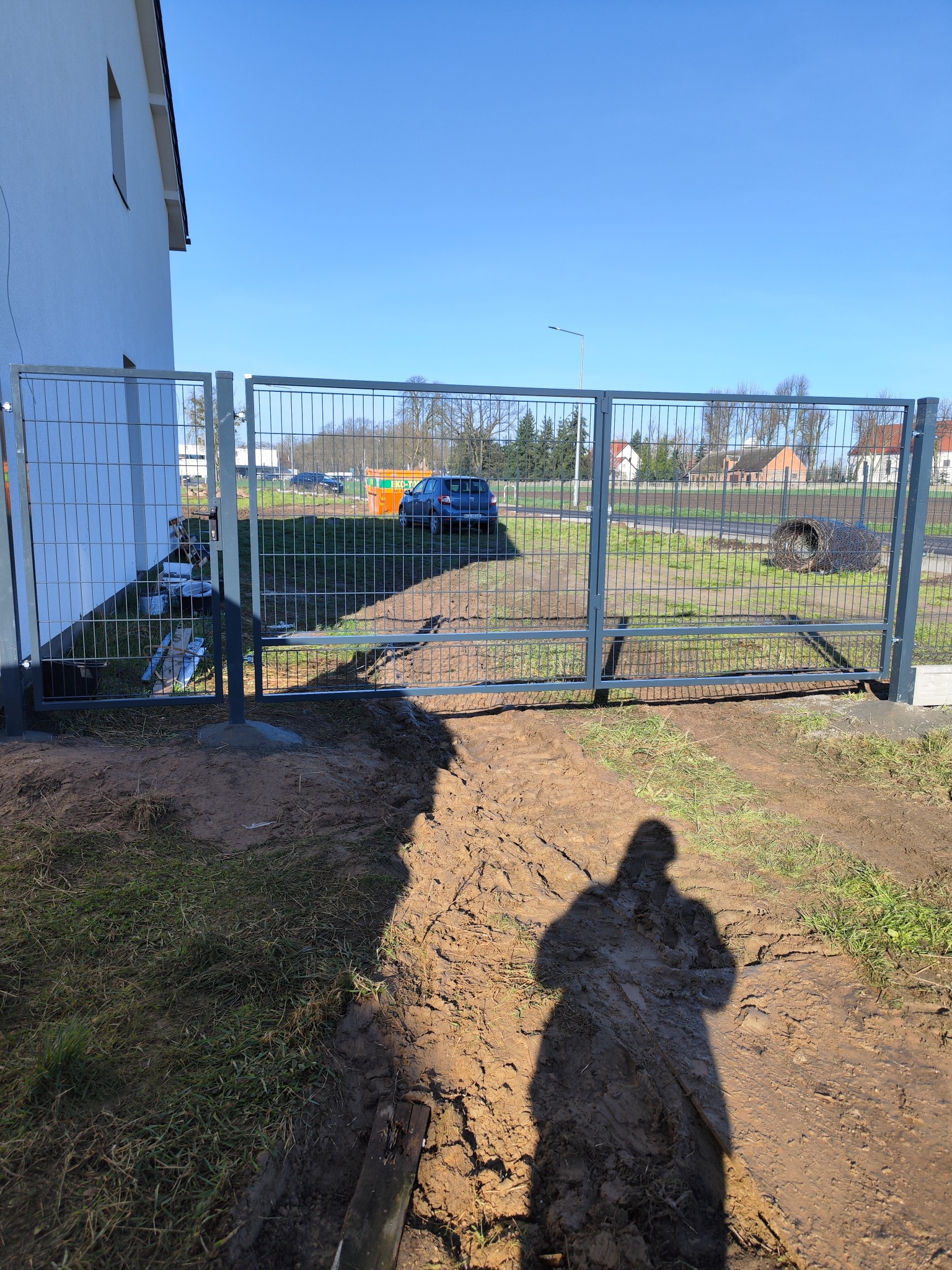 Szara brama panelowa z widoczną posesją w tle, cień osoby fotografującej na pierwszym planie.