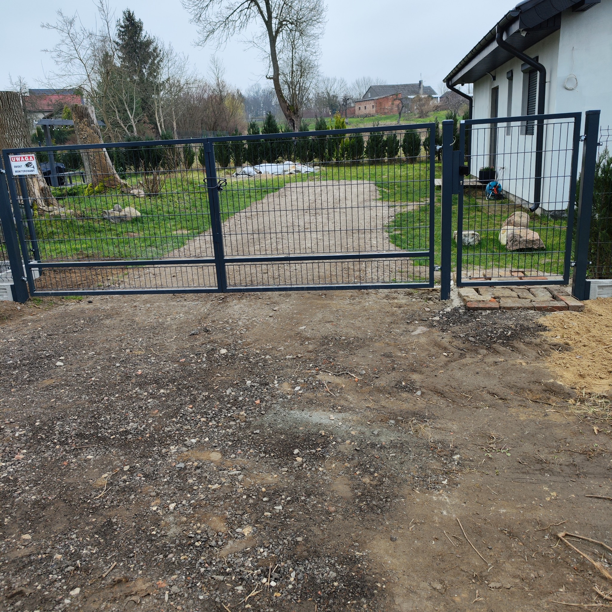 Nowoczesna posesja z dwuskrzydłową bramą panelową w kolorze antracytowym, widok na żwirową drogę dojazdową i dom z białymi ścianami.