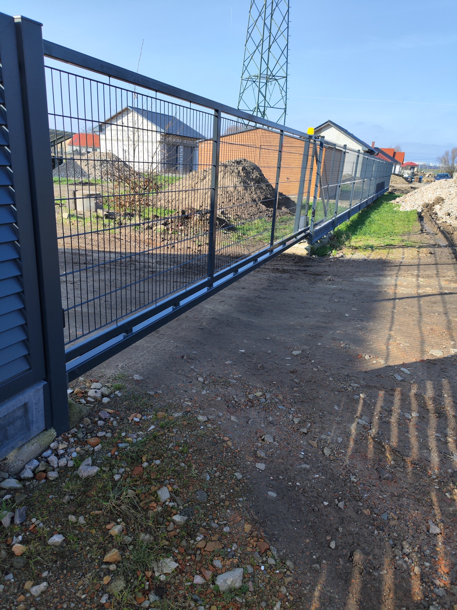 Przesuwna brama z paneli ogrodzeniowych z automatyką, widoczne domy w tle i słup wysokiego napięcia.