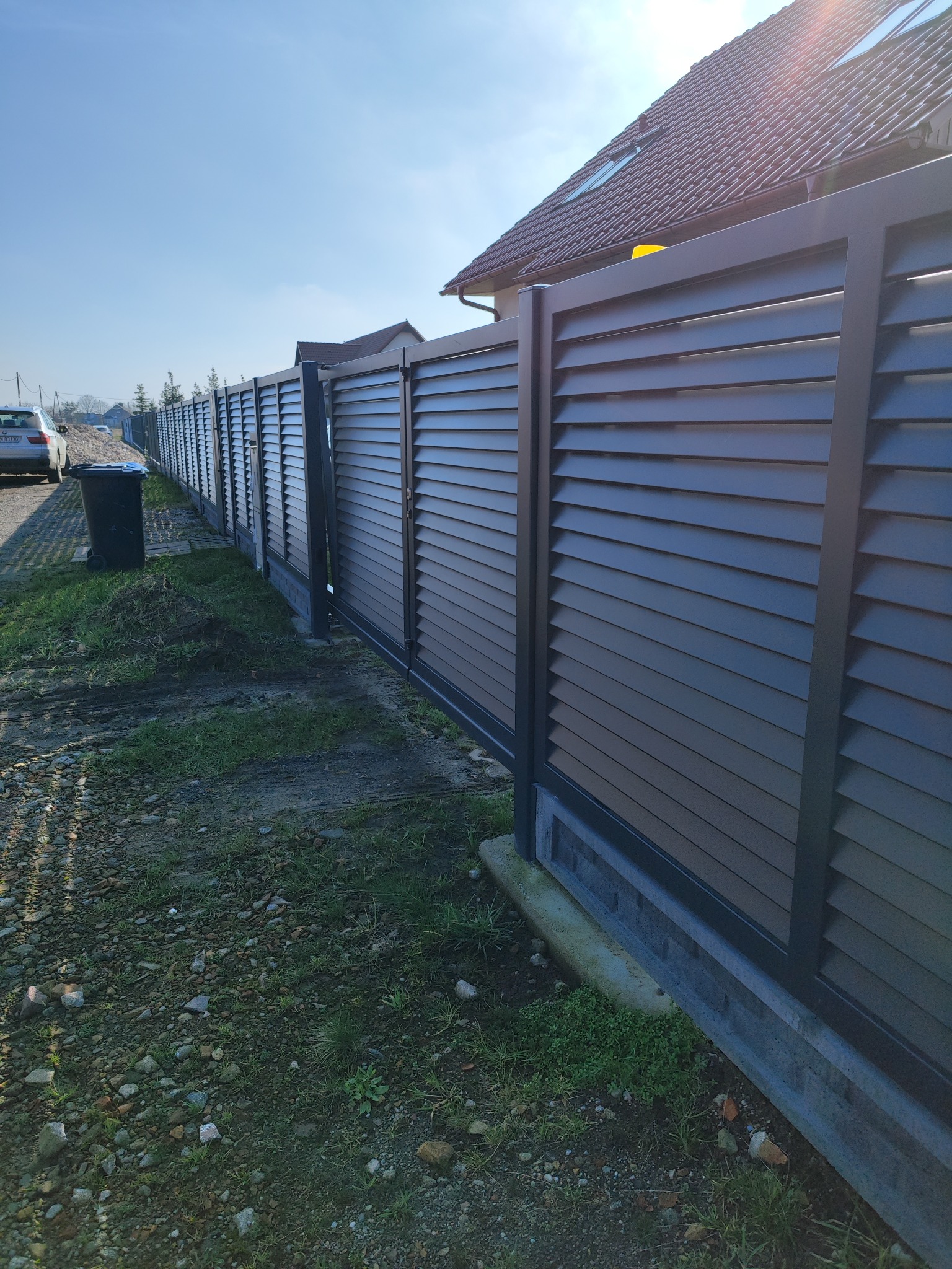 Długa, nowoczesna, pozioma, szara przesłona z metalu, tworząca ogrodzenie posesji, widoczna z zewnątrz, z domem w tle.