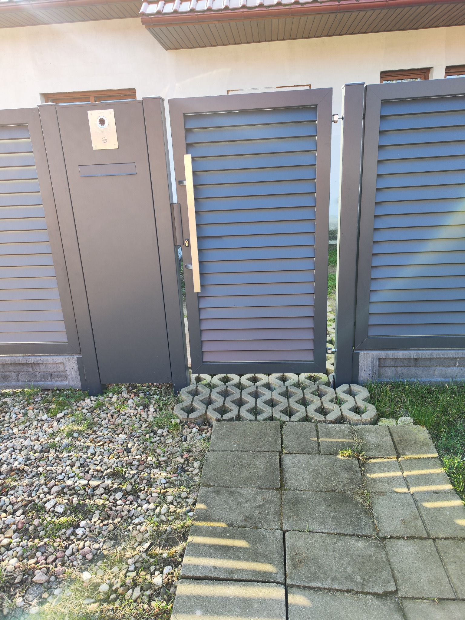 Nowoczesna, szara brama z poziomymi lamelami i furtka z wideodomofonem, zamontowana na posesji z kamiennym podłożem i betonowymi płytami.