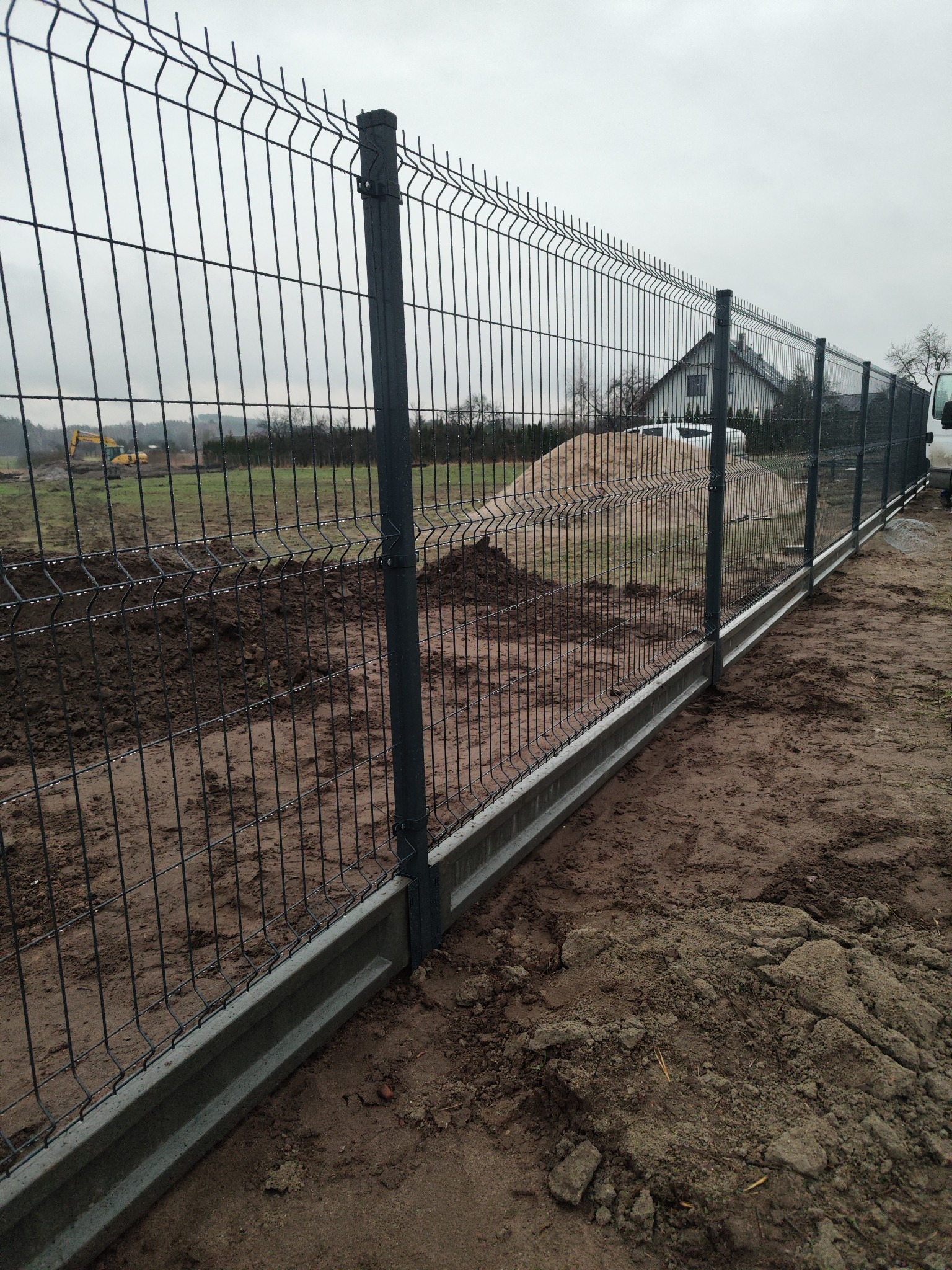 Nowo zainstalowane, antracytowe ogrodzenie z paneli z podmurówką betonową na działce w trakcie przygotowania, w tle dom i koparka.