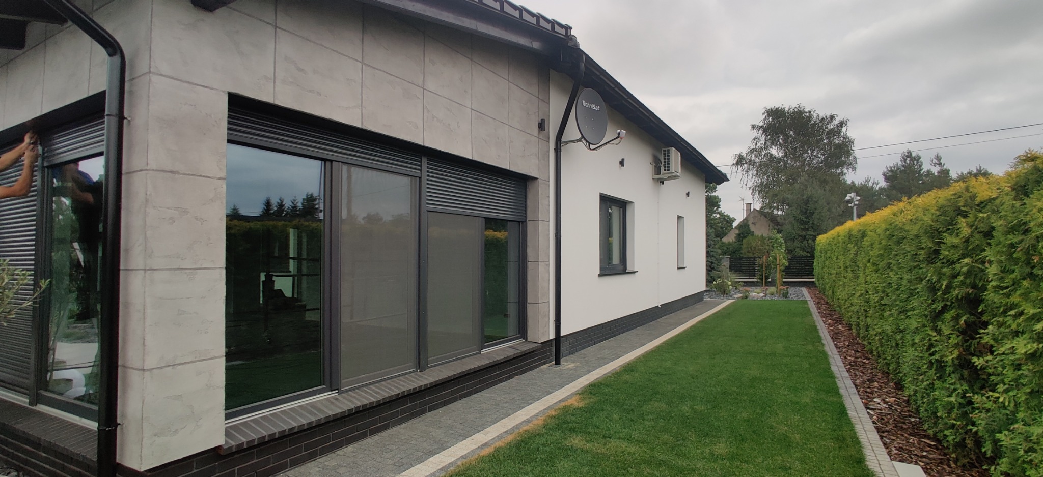 Elewacja domu jednorodzinnego z zamontowanymi szarymi roletami zewnętrznymi, częściowo opuszczonymi, widoczne ręce montera przy oknie, obok zadbany trawnik i żywopłot.