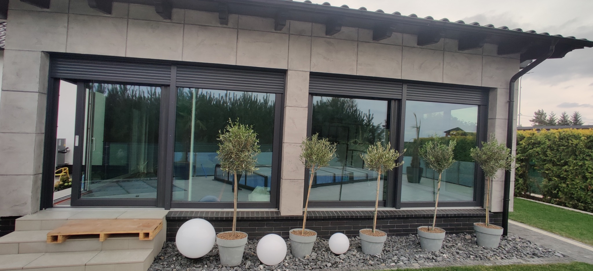 Nowoczesny dom z szarymi oknami, częściowo zasłoniętymi zewnętrznymi roletami, ozdobiony drzewkami w donicach i kulistymi lampami na tle zielonego trawnika.