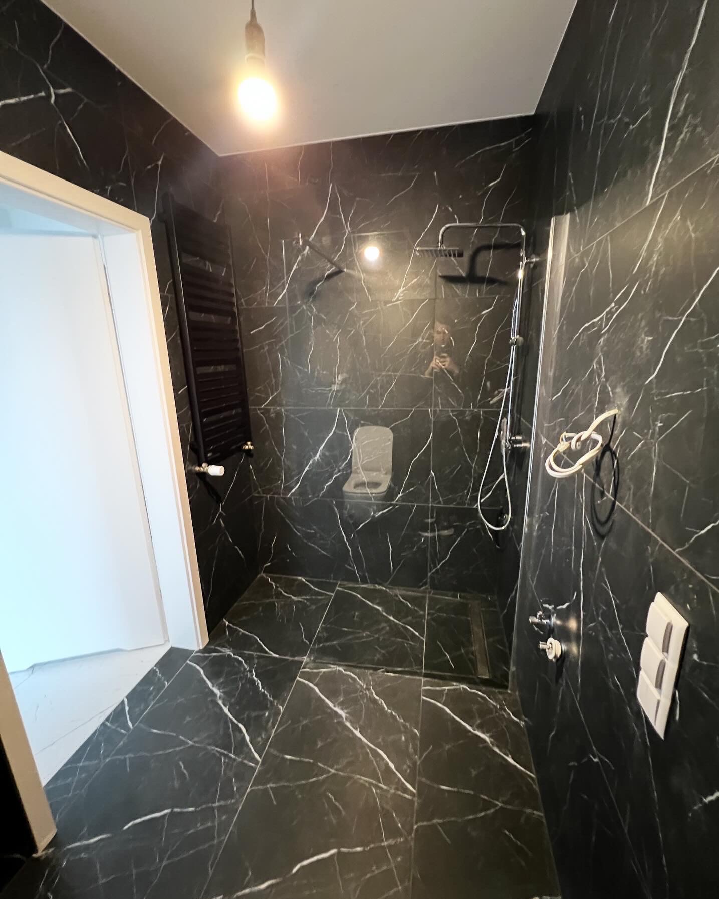 Wykończona łazienka z czarnymi płytkami imitującymi marmur, kabina prysznicowa typu walk-in z odpływem liniowym, grzejnik drabinkowy w kolorze czarnym, biała armatura i oświetlenie punktowe.