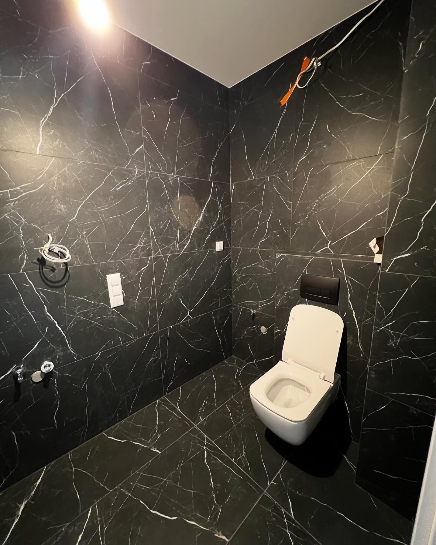 Nowoczesna łazienka z czarnymi płytkami imitującymi marmur, wisząca toaleta z czarnym przyciskiem spłukującym, widoczne przyłącza wodne i niedokończone podłączenie elektryczne.