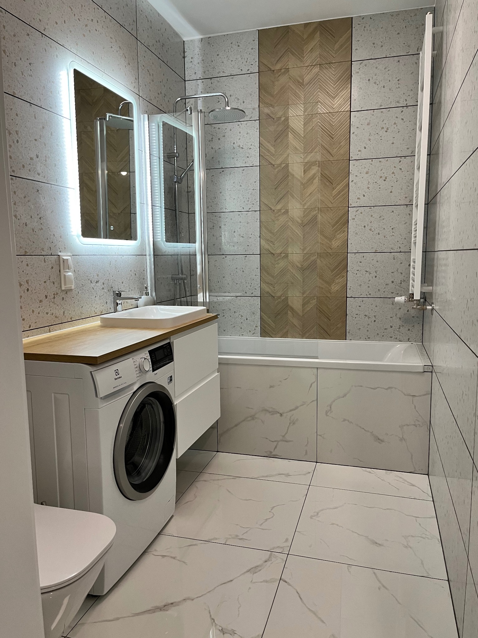 Nowoczesna łazienka z pralką pod blatem, jasnymi płytkami imitującymi kamień, akcentem z płytek drewnopodobnych ułożonych w jodełkę, podświetlanym lustrem i kabiną prysznicową.