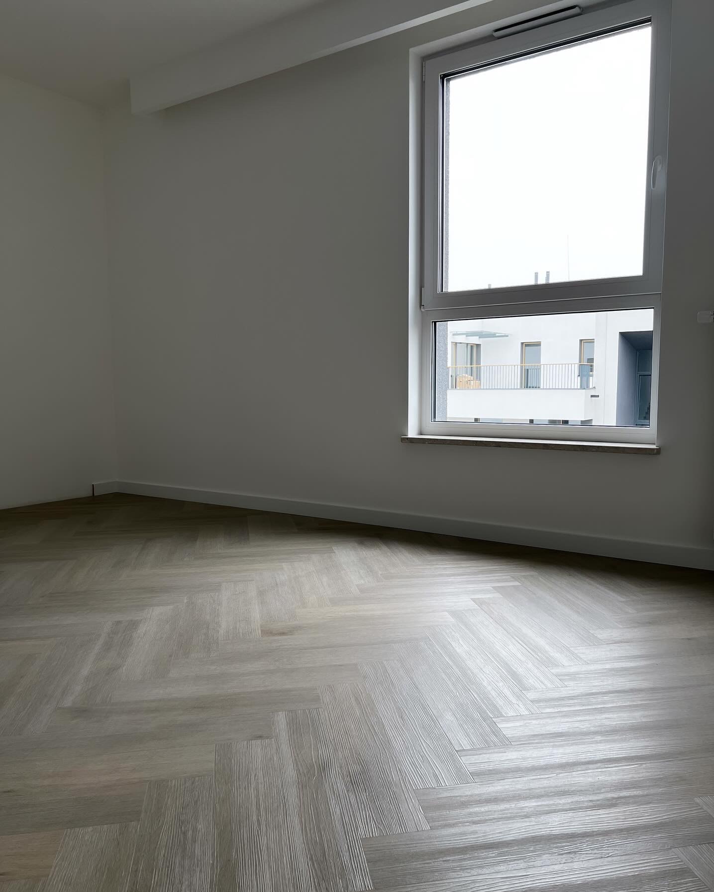 Jasnoszara podłoga ułożona w jodełkę w pustym pokoju z dużym oknem, widok na nowoczesny budynek.