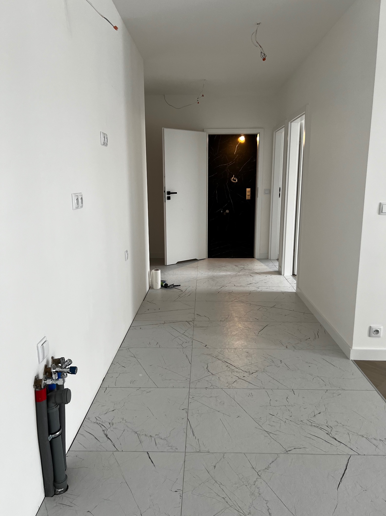 Korytarz w trakcie prac wykończeniowych: białe ściany z wystającymi kablami, szare płytki imitujące marmur na podłodze, instalacja wodno-kanalizacyjna przy ścianie, otwarte drzwi do łazienki...