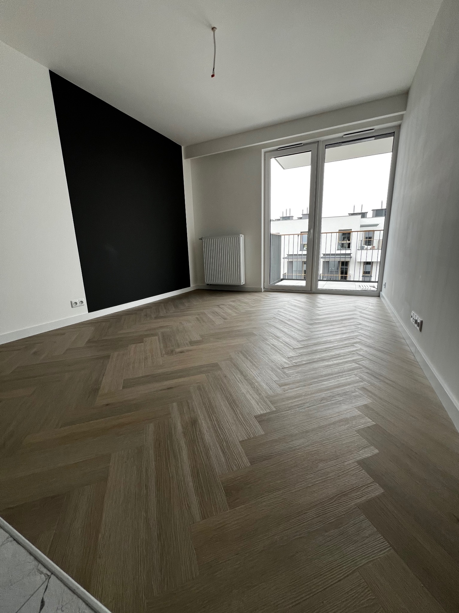 Nowoczesne wnętrze z parkietem ułożonym w jodełkę, czarną ścianą kontrastującą z bielą, dużym oknem balkonowym i grzejnikiem.