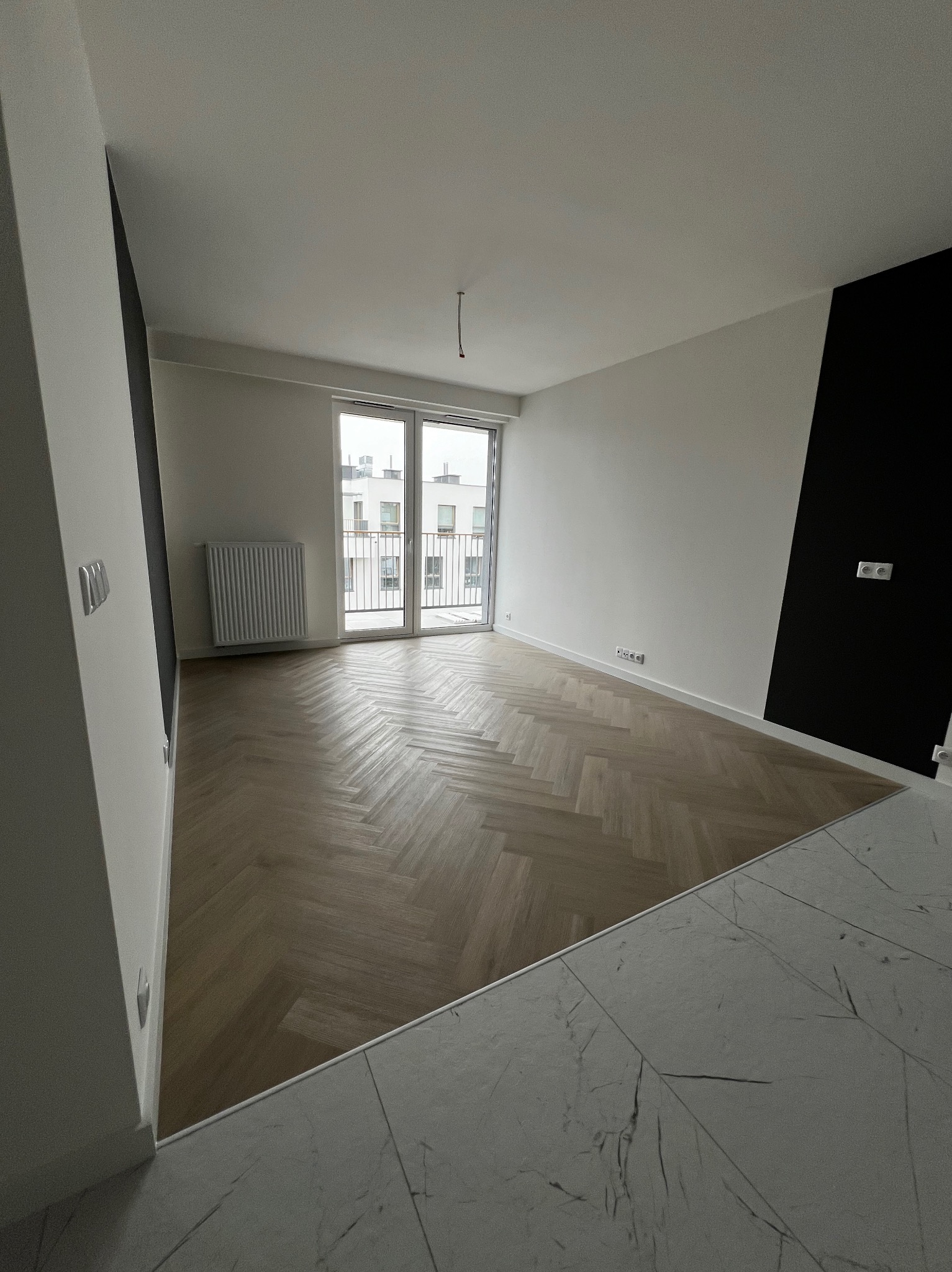 Pusty pokój z parkietem ułożonym w jodełkę, białymi ścianami z czarnym akcentem, grzejnikiem i balkonem z widokiem na sąsiedni budynek.
