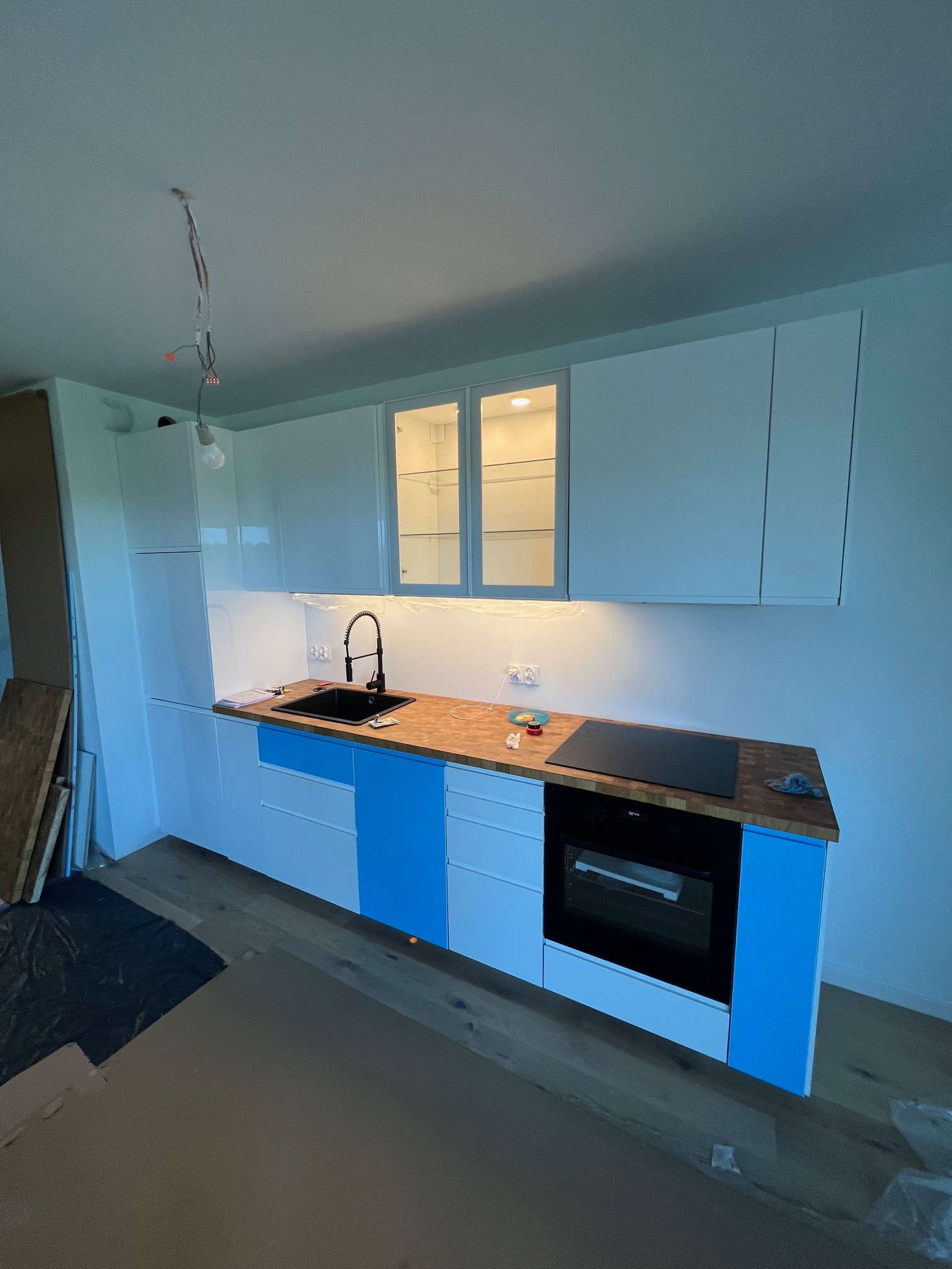 Częściowo zamontowana, nowoczesna kuchnia z białymi szafkami górnymi z podświetleniem i niebieskimi szafkami dolnymi, drewnianym blatem, czarnym zlewem i płytą indukcyjną, widoczne niedokończone...
