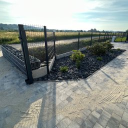 Brukowanie Łukasz Kosz - Nowoczesne ogrodzenie panelowe z gabionami wypełnionymi czarnym kamieniem, obok wybrukowany chodnik z piaskiem i widok na pole w tle.