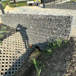 Brukowanie Łukasz Kosz - Gabionowy murek oporowy z odpływem wody, ażurowe płyty betonowe na skarpie, widoczne elementy drenażu i roślinność.