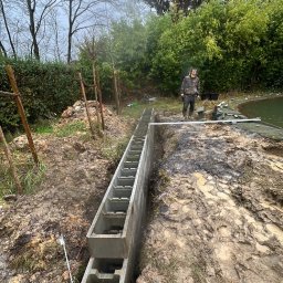 Brukowanie Łukasz Kosz - Budowa fundamentu pod ogrodzenie z użyciem prefabrykowanych betonowych elementów w wykopie, widoczny pracownik w oddali i błotniste otoczenie.