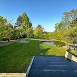 Brukowanie Łukasz Kosz - Widok na zadbany ogród z oczkiem wodnym i drewnianym pomostem, widoczne schody z ciemnych płyt prowadzące w dół, równo przystrzyżony trawnik, drzewa i krzewy w tle.