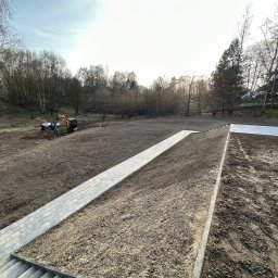 Brukowanie Łukasz Kosz - Przygotowanie terenu pod budowę: widok z góry na częściowo ułożony chodnik z kostki brukowej, minikoparka i pracownik w oddali, świeżo nawieziona ziemia, drzewa w tle.