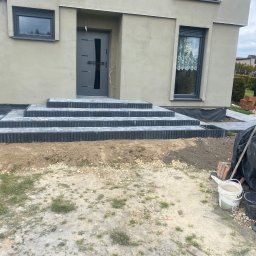 Brukowanie Łukasz Kosz - Ułożone schody wejściowe z kostki brukowej w kolorze grafitowym, z widocznymi obrzeżami, prowadzące do nowoczesnego domu z szarą elewacją i stolarką okienną, w tle widoczne są narzędzia i materiały...