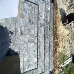 Brukowanie Łukasz Kosz - Widok z góry na świeżo ułożoną, szarą kostkę brukową tworzącą schody i chodnik, z widocznymi elementami mocującymi na krawędziach i obrzeżem z ziemi oraz fragmentem buta osoby fotografującej...