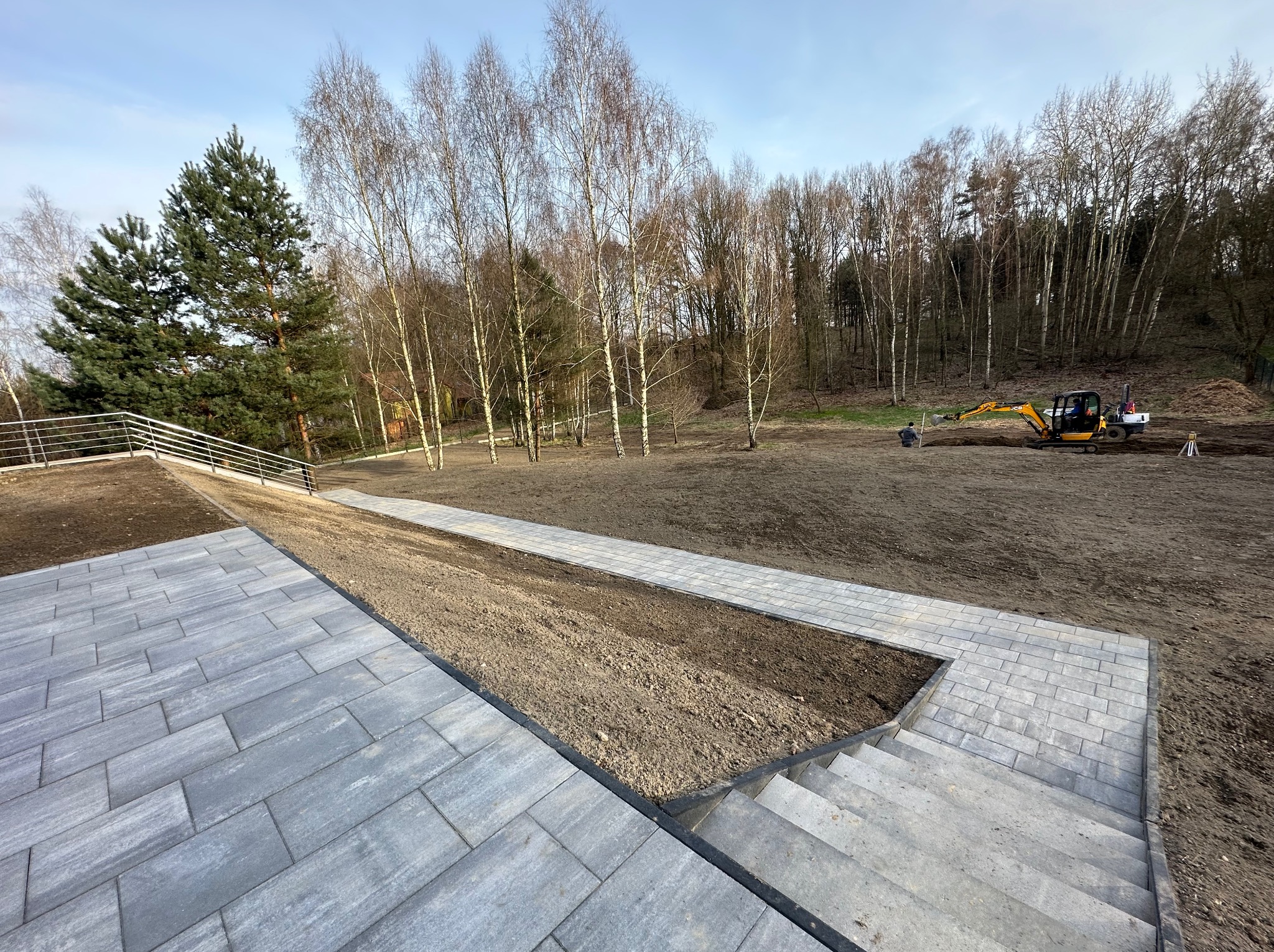 Układanie kostki brukowej na posesji z wybrukowanymi schodami i ścieżką, w tle krajobraz z drzewami i pracujący mały żółty koparka.