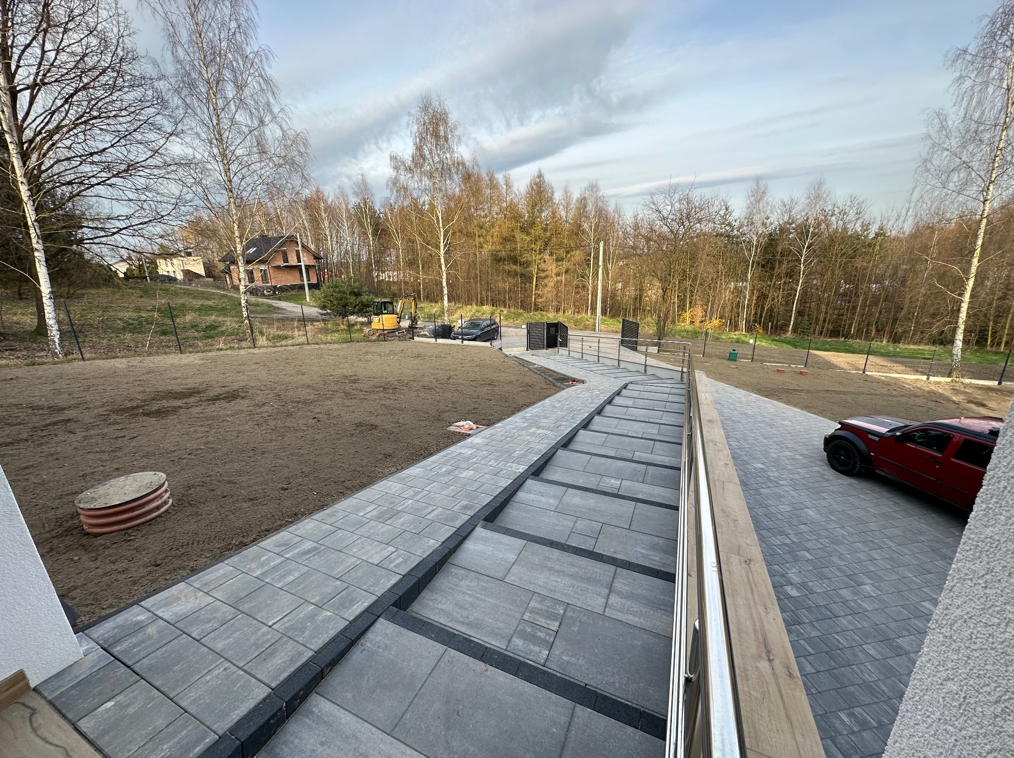 Szerokie, schodkowe podejście z szarych płyt betonowych prowadzące w dół, z metalową barierką po prawej stronie; w tle dom, teren przygotowany pod trawnik i zaparkowane samochody, w tym czerwony SUV.