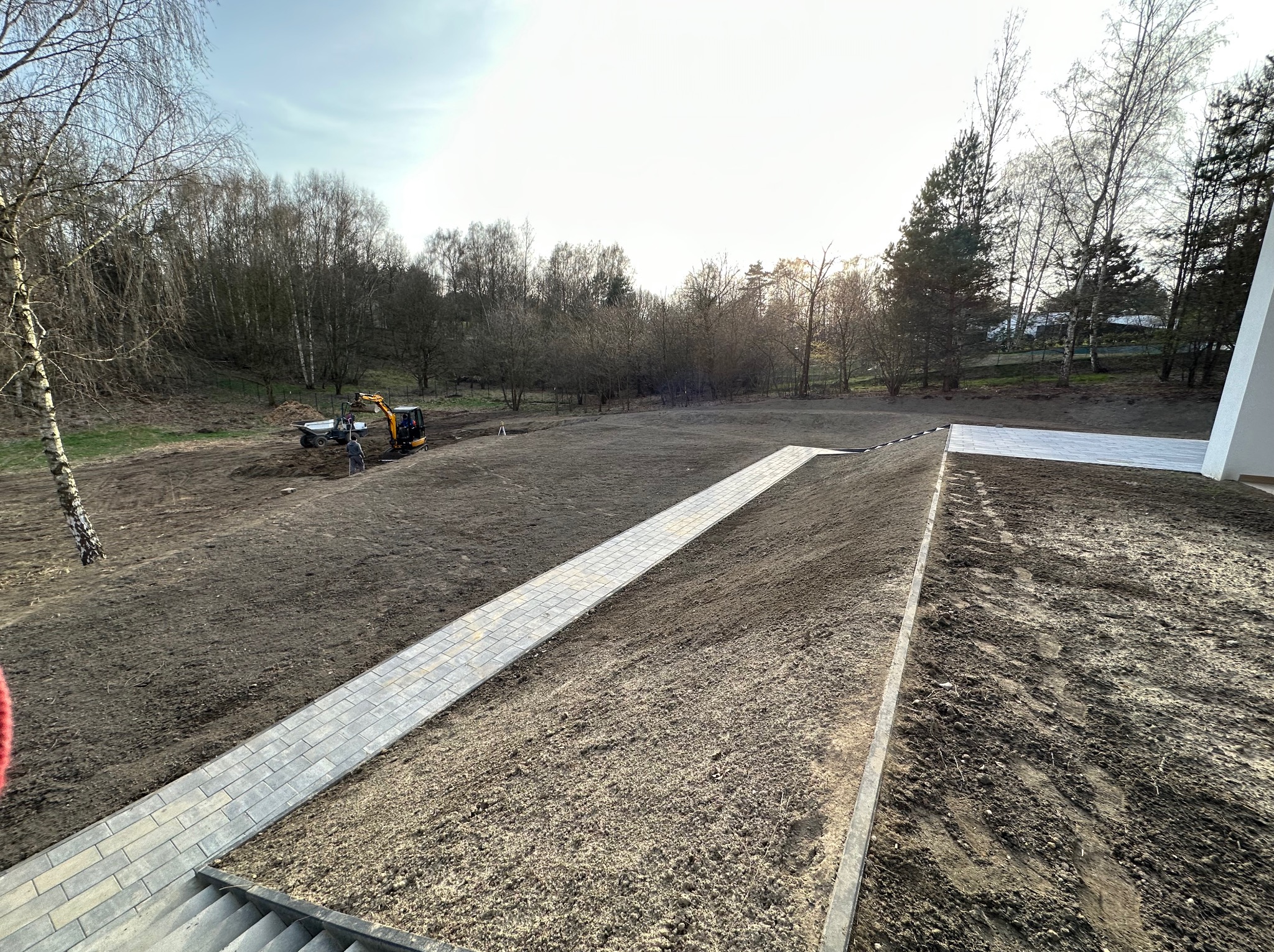 Przygotowanie terenu pod budowę: widok z góry na częściowo ułożony chodnik z kostki brukowej, minikoparka i pracownik w oddali, świeżo nawieziona ziemia, drzewa w tle.