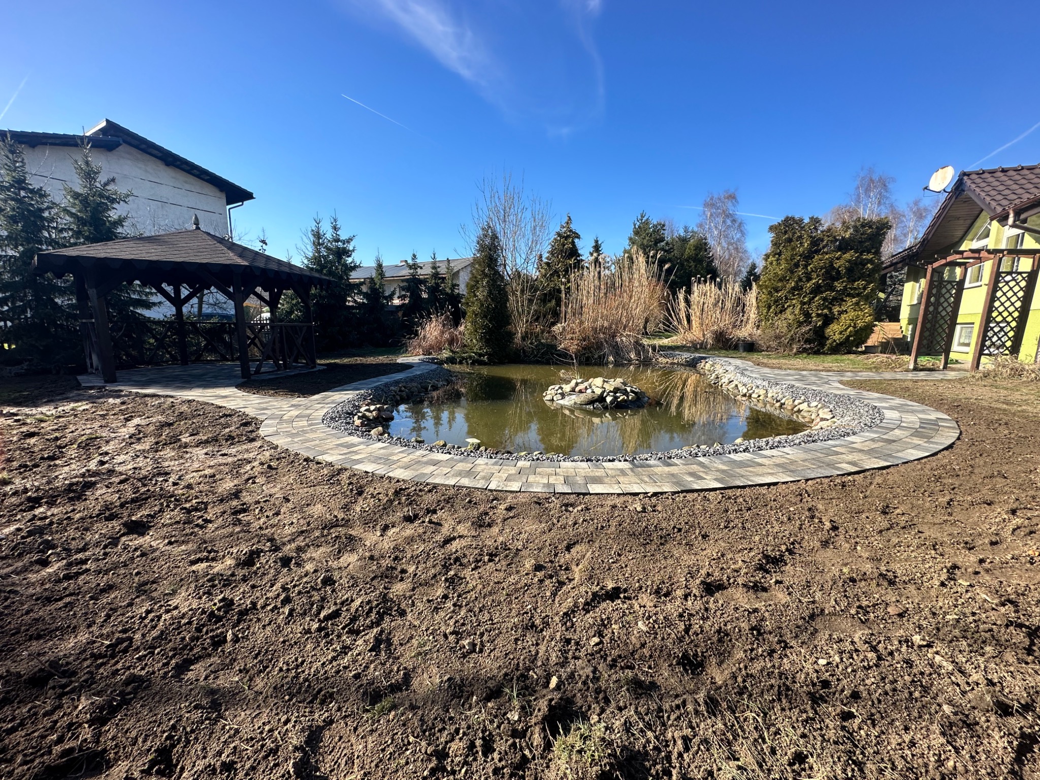 Oczko wodne otoczone kostką brukową i kamieniami w ogrodzie, z widoczną altaną w tle oraz przygotowanym podłożem pod trawnik.