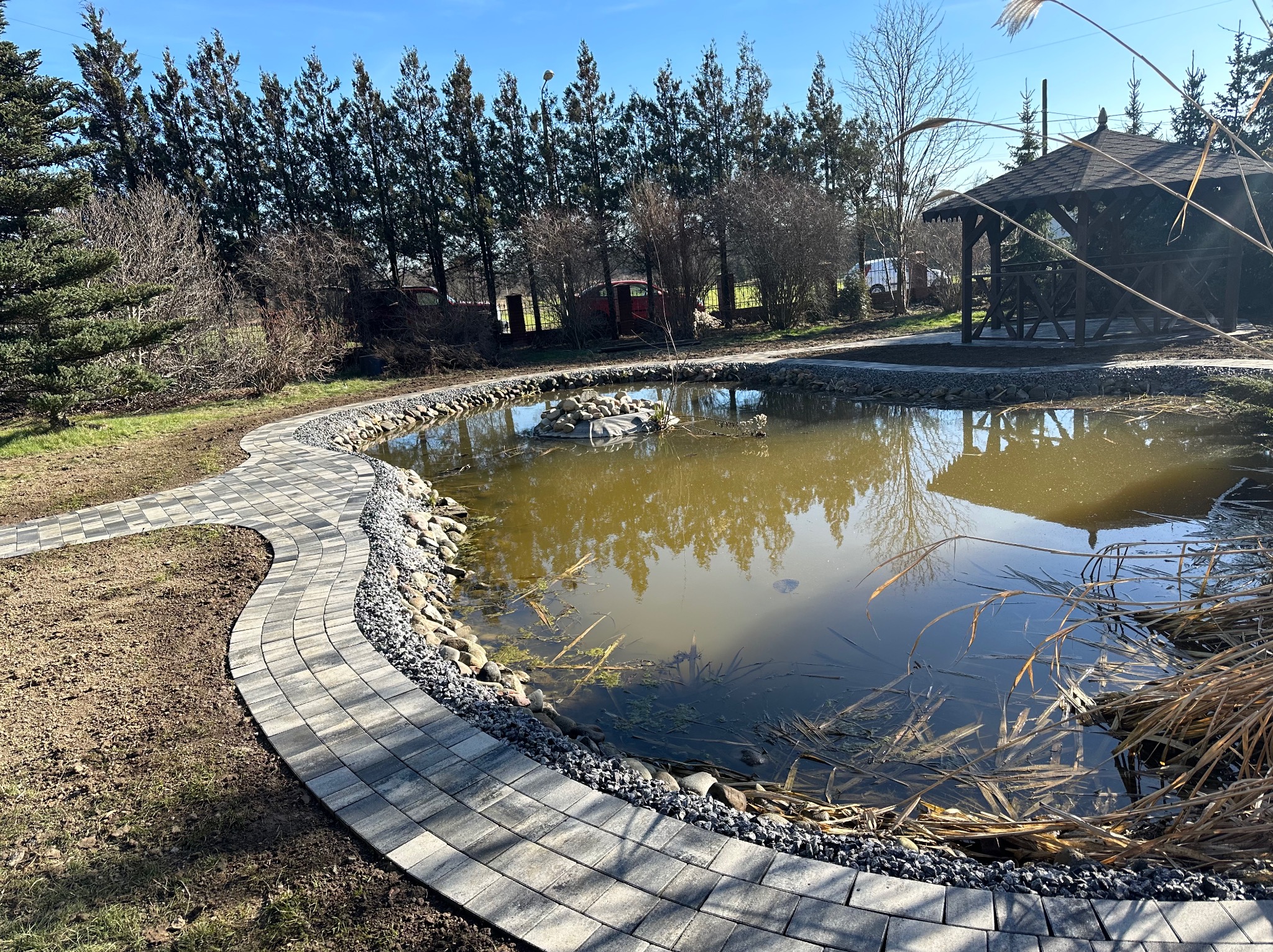 Ogród z oczkiem wodnym obłożonym kamieniami i ścieżką z szarej kostki brukowej, w tle altana i rząd wysokich drzew iglastych.