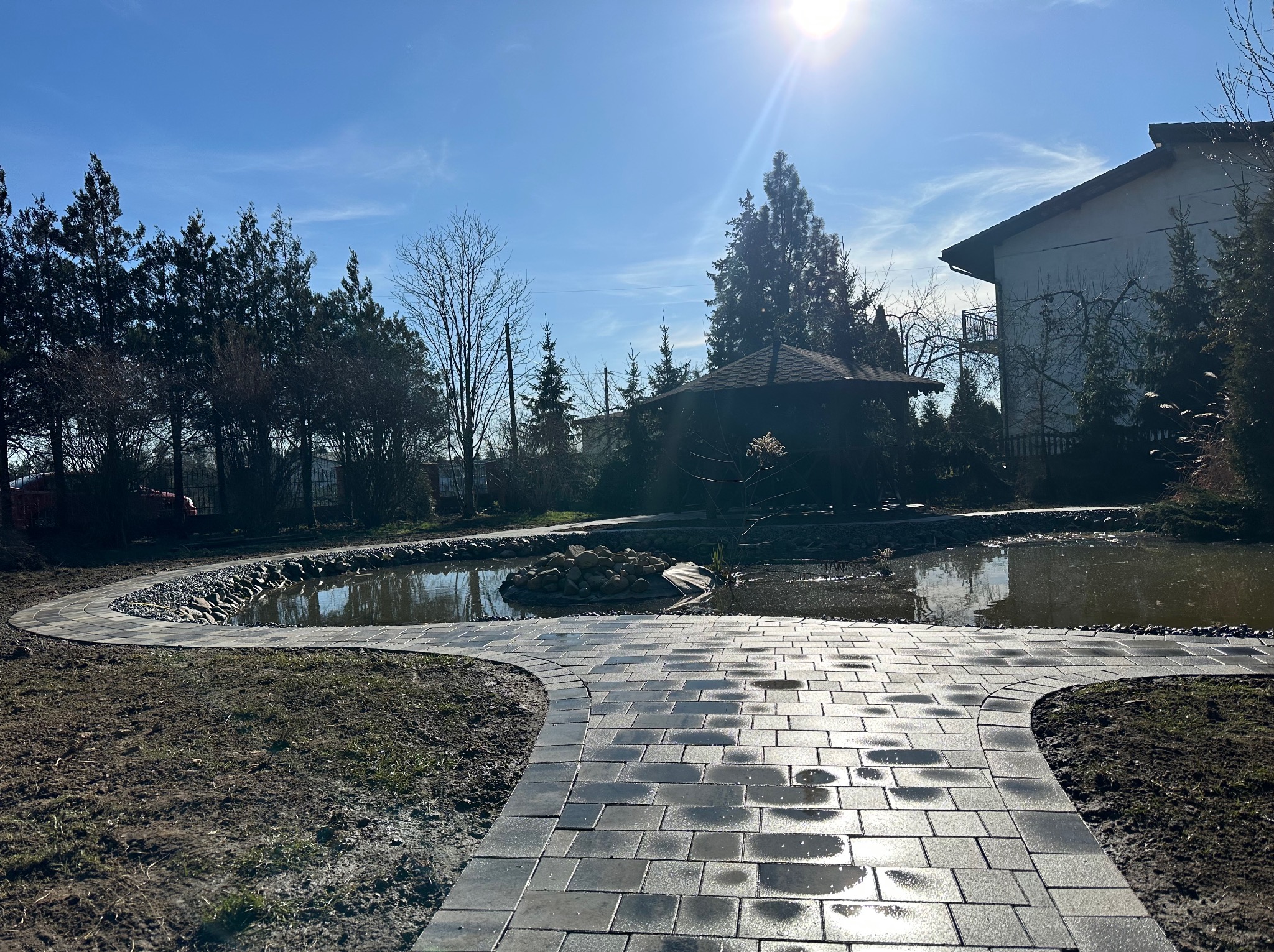 Ścieżka z kostki brukowej rozwidlająca się w kierunku stawu z kamieniami i altany ogrodowej w słoneczny dzień.