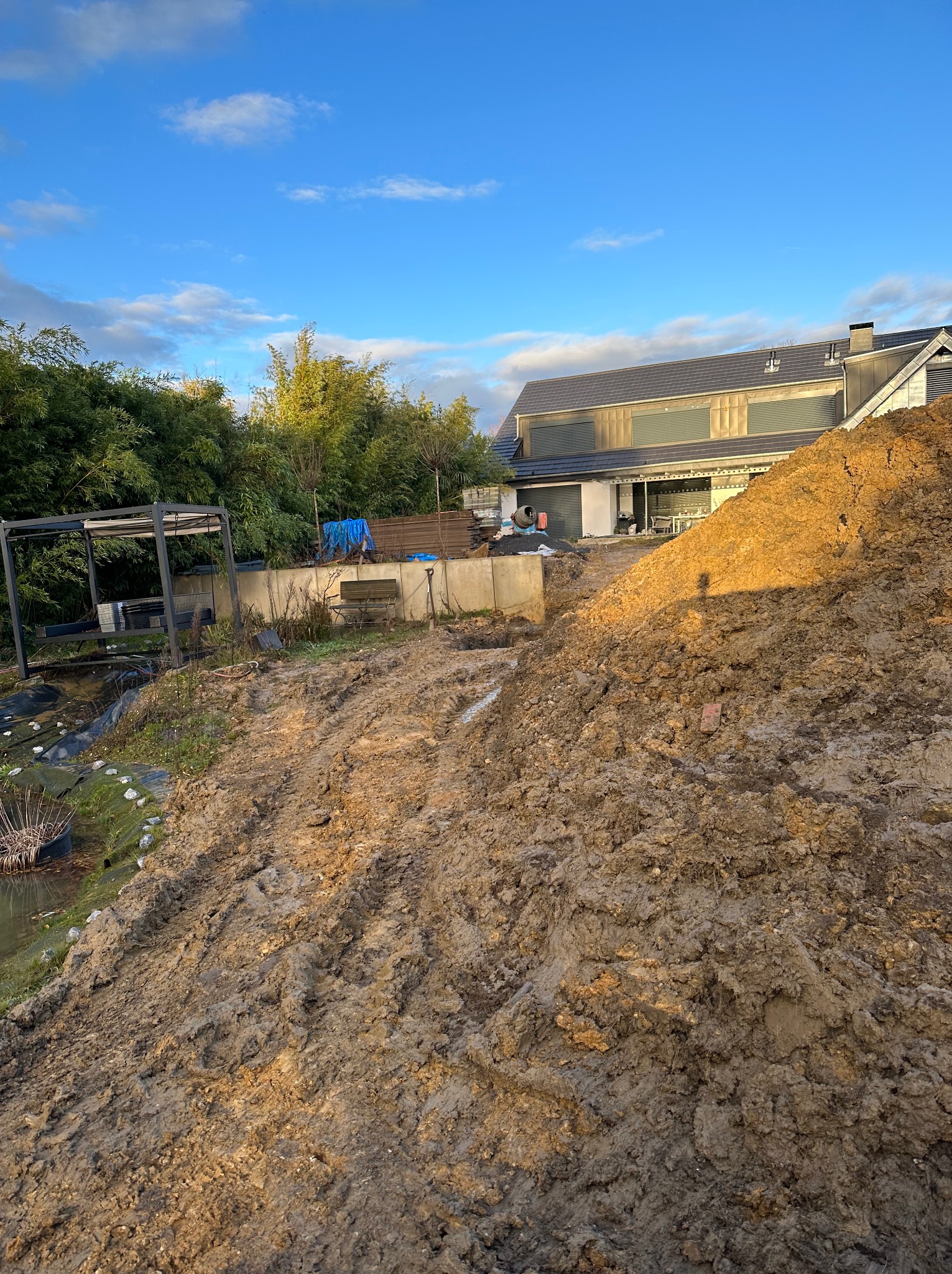 Stos ziemi z widocznymi śladami opon w ogrodzie przed nowoczesnym domem z szarą elewacją, w tle zieleń drzew i błękitne niebo.