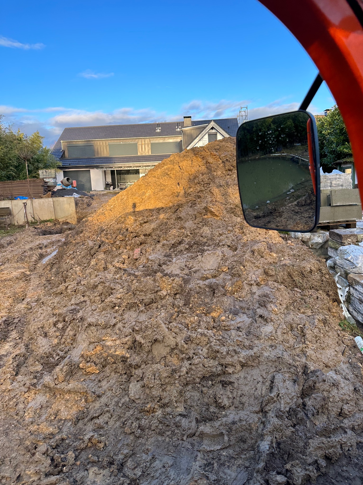 Góra ziemi z widokiem z kabiny maszyny budowlanej, odbicie w lusterku widoczne, w tle dom w trakcie budowy.