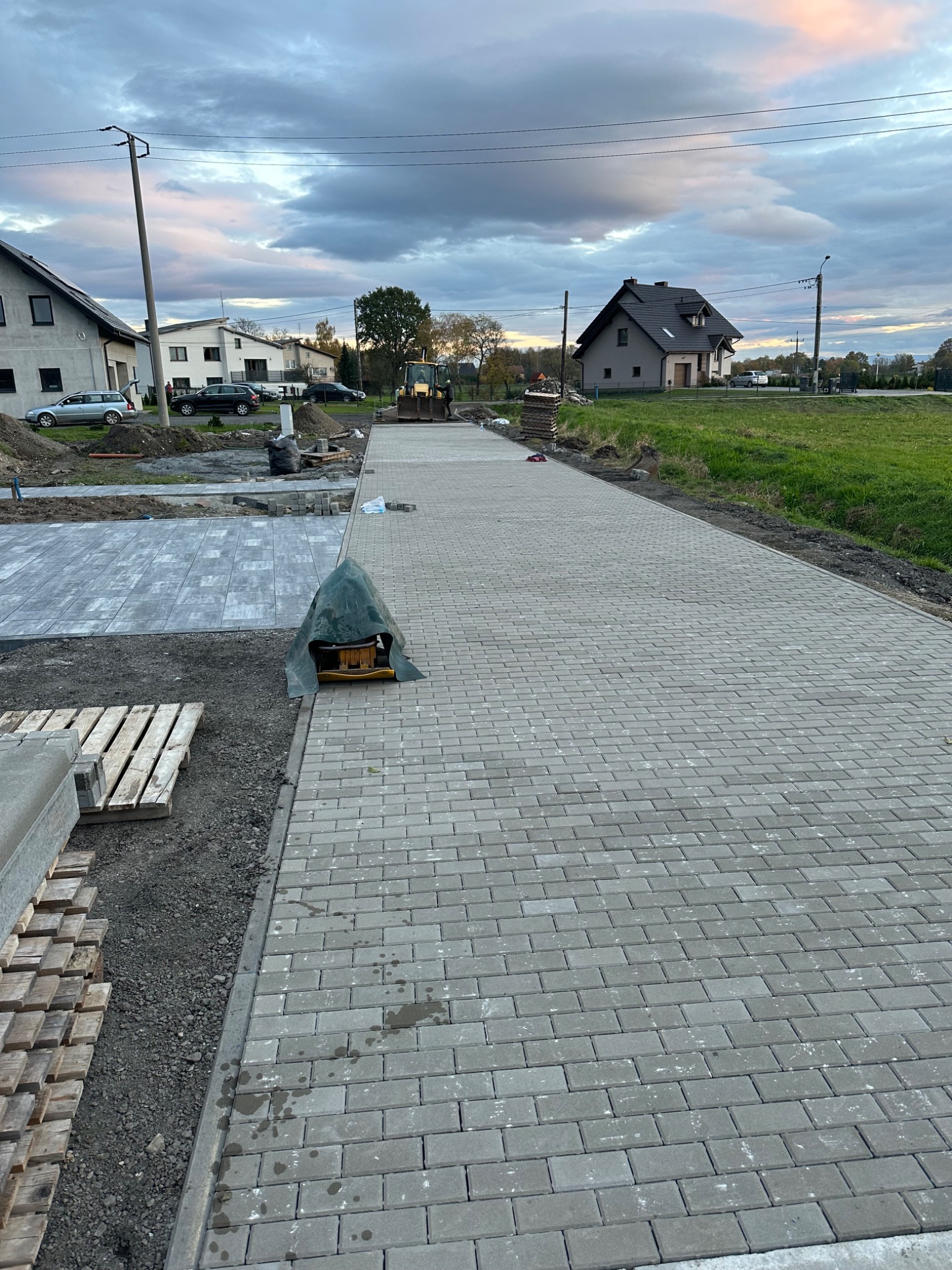 Szeroki podjazd z szarej kostki brukowej w trakcie budowy, widoczna ubijarka, palety z kostką i koparka w tle, domy jednorodzinne w sąsiedztwie.