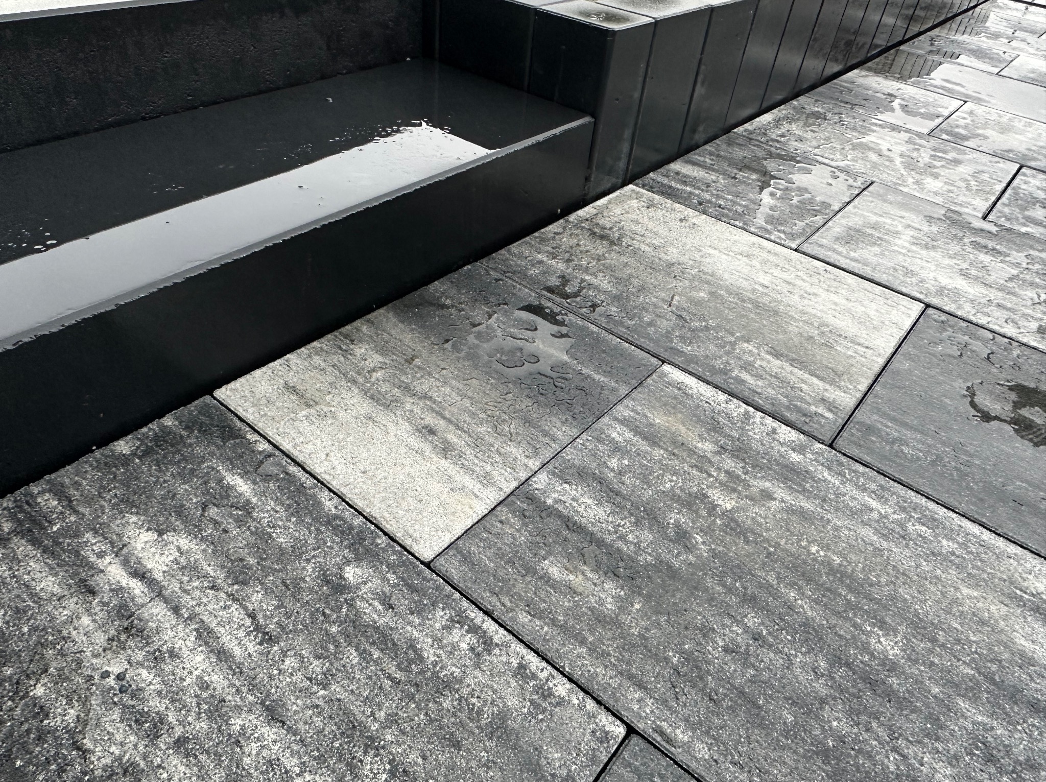 Szara kostka brukowa ułożona pod kątem, mokra od deszczu, widoczne krople wody na powierzchni, obok betonowy element architektoniczny z wodą.