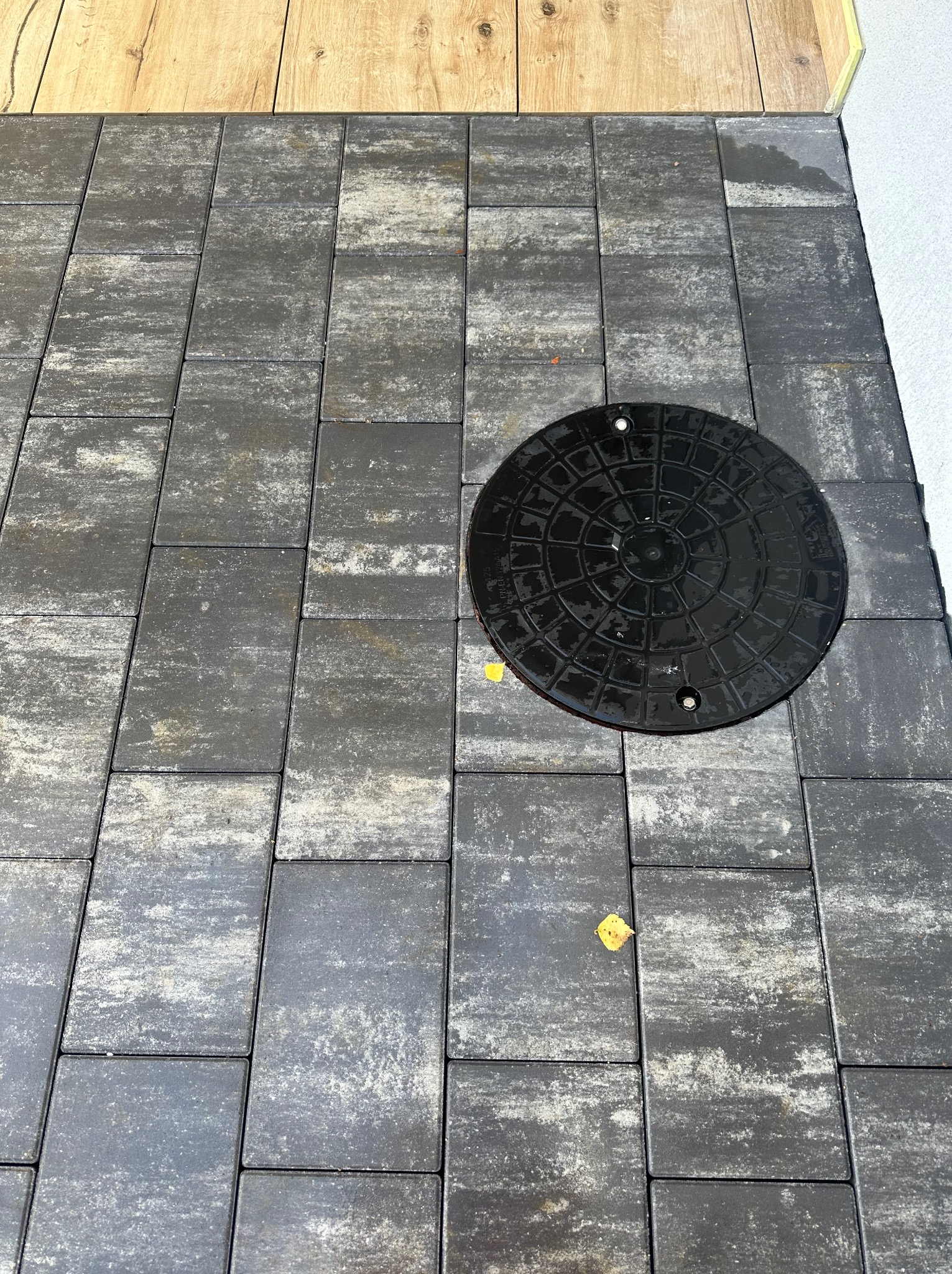 Ułożona kostka brukowa w odcieniach szarości z widoczną studzienką kanalizacyjną oraz fragmentem drewnianej podłogi lub tarasu.