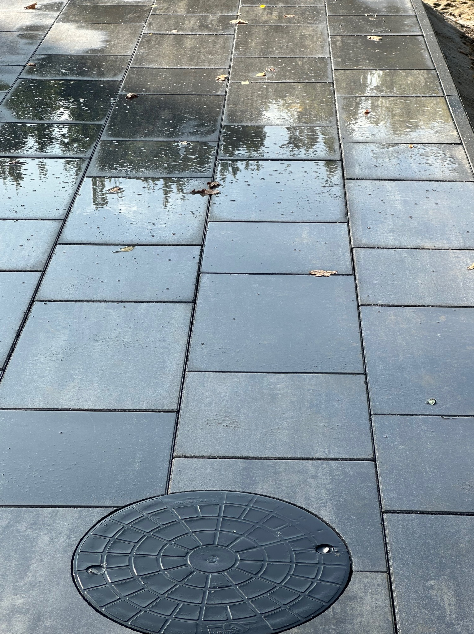 Świeżo ułożona, mokra nawierzchnia z szarych, prostokątnych płyt betonowych z widoczną studzienką kanalizacyjną w dolnej części kadru i kilkoma opadłymi liśćmi na powierzchni.