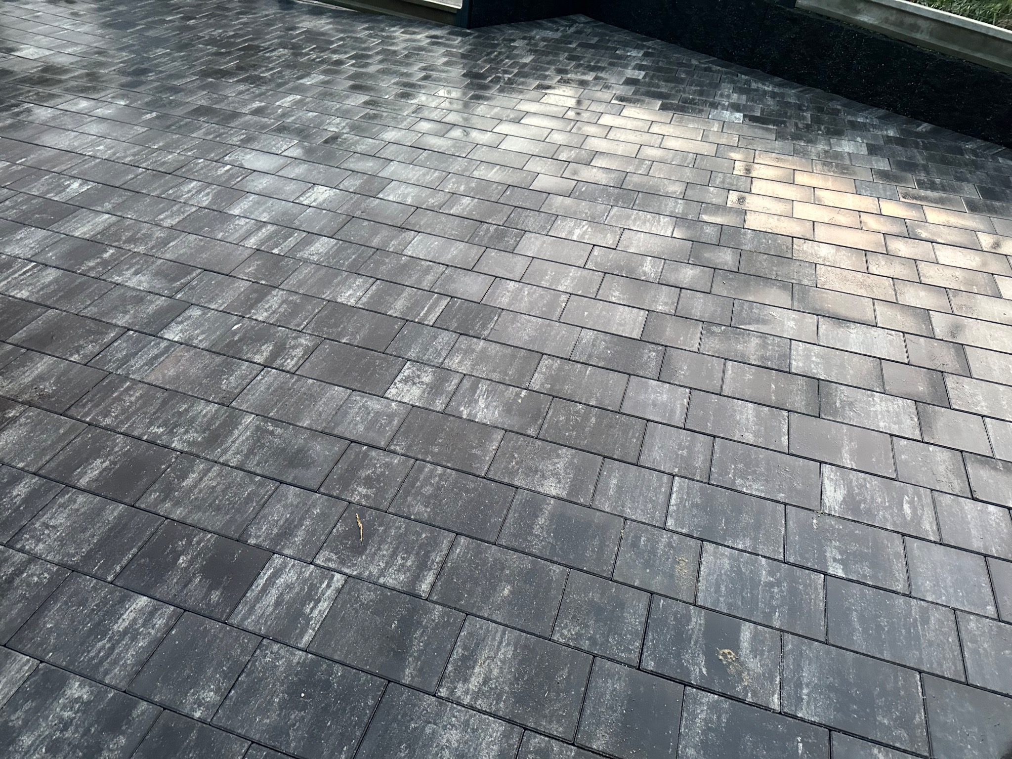 Ułożona, szara kostka brukowa w formacie prostokątów, lekko wilgotna, z widocznymi śladami użytkowania i pojedynczym, małym listkiem, ułożona pod kątem z widocznym fragmentem budynku w tle.
