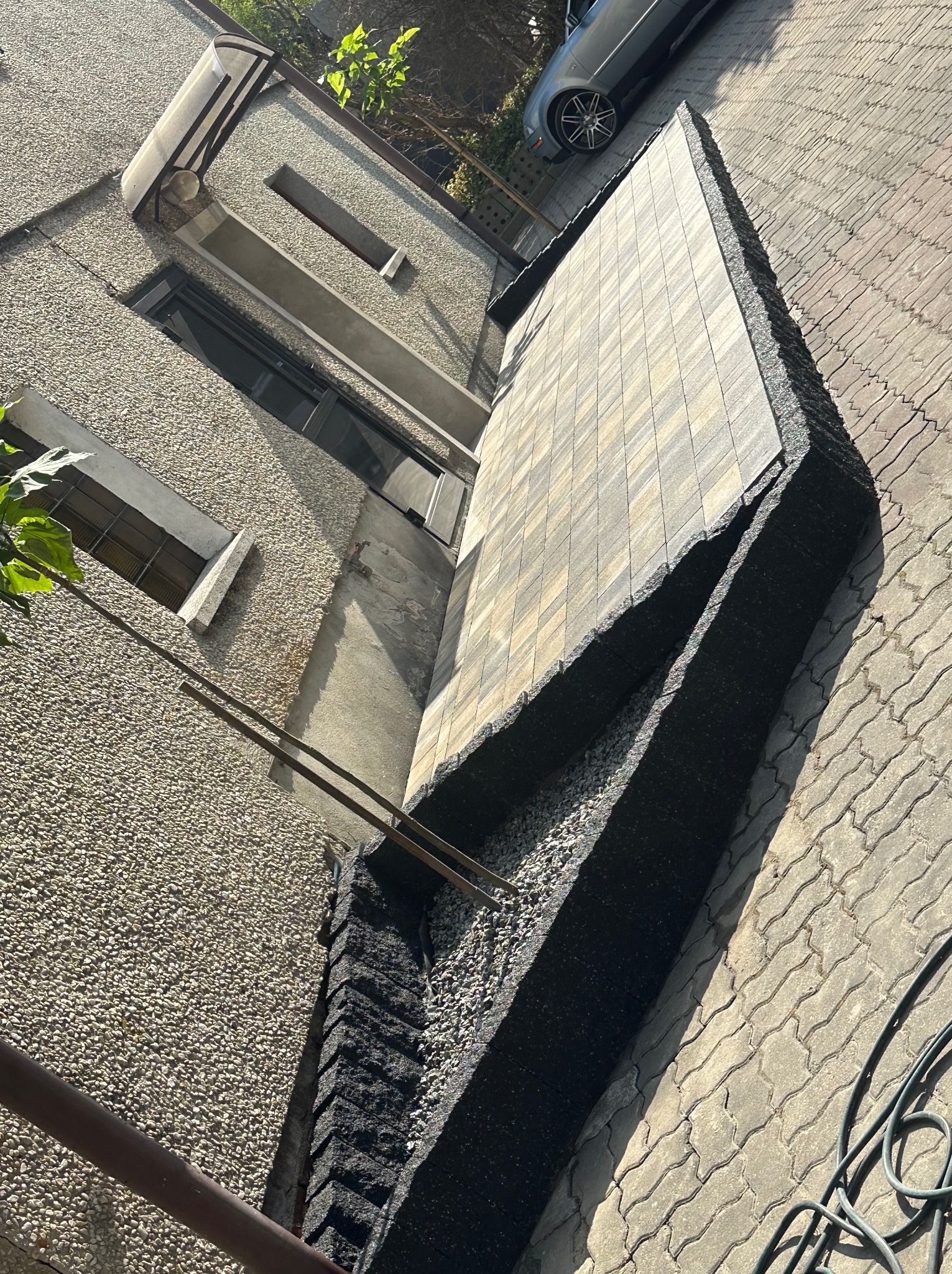 Fragment układanej kostki brukowej przy ścianie budynku, widoczne warstwy podbudowy i krawężniki, obok stara kostka brukowa i zaparkowany samochód.