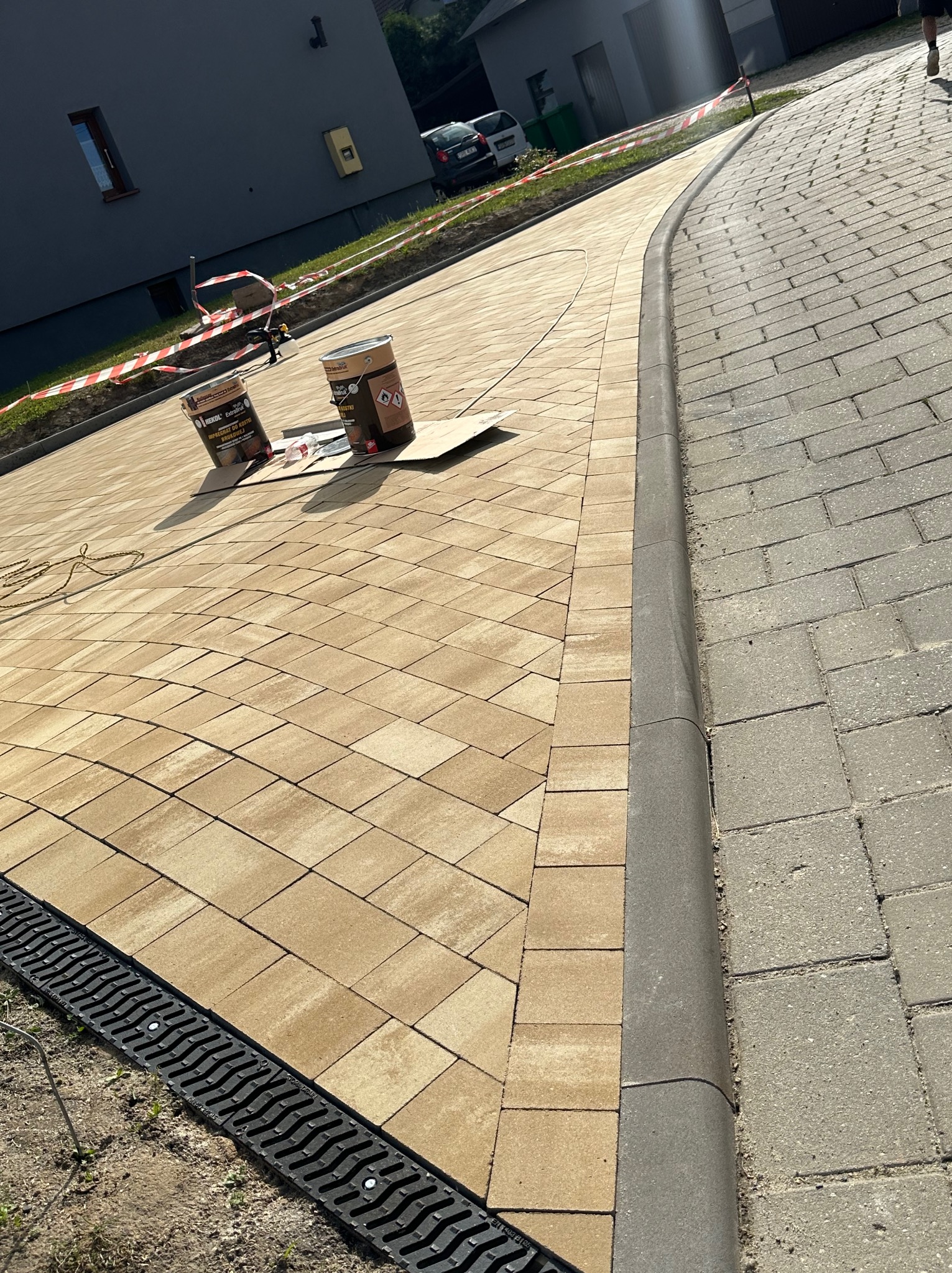 Nowo ułożona, beżowa kostka brukowa na podjeździe, z widocznym odpływem liniowym i krawężnikiem, zaimpregnowana preparatem z dwóch puszek stojących na kartonie, odseparowana taśmą ostrzegawczą...