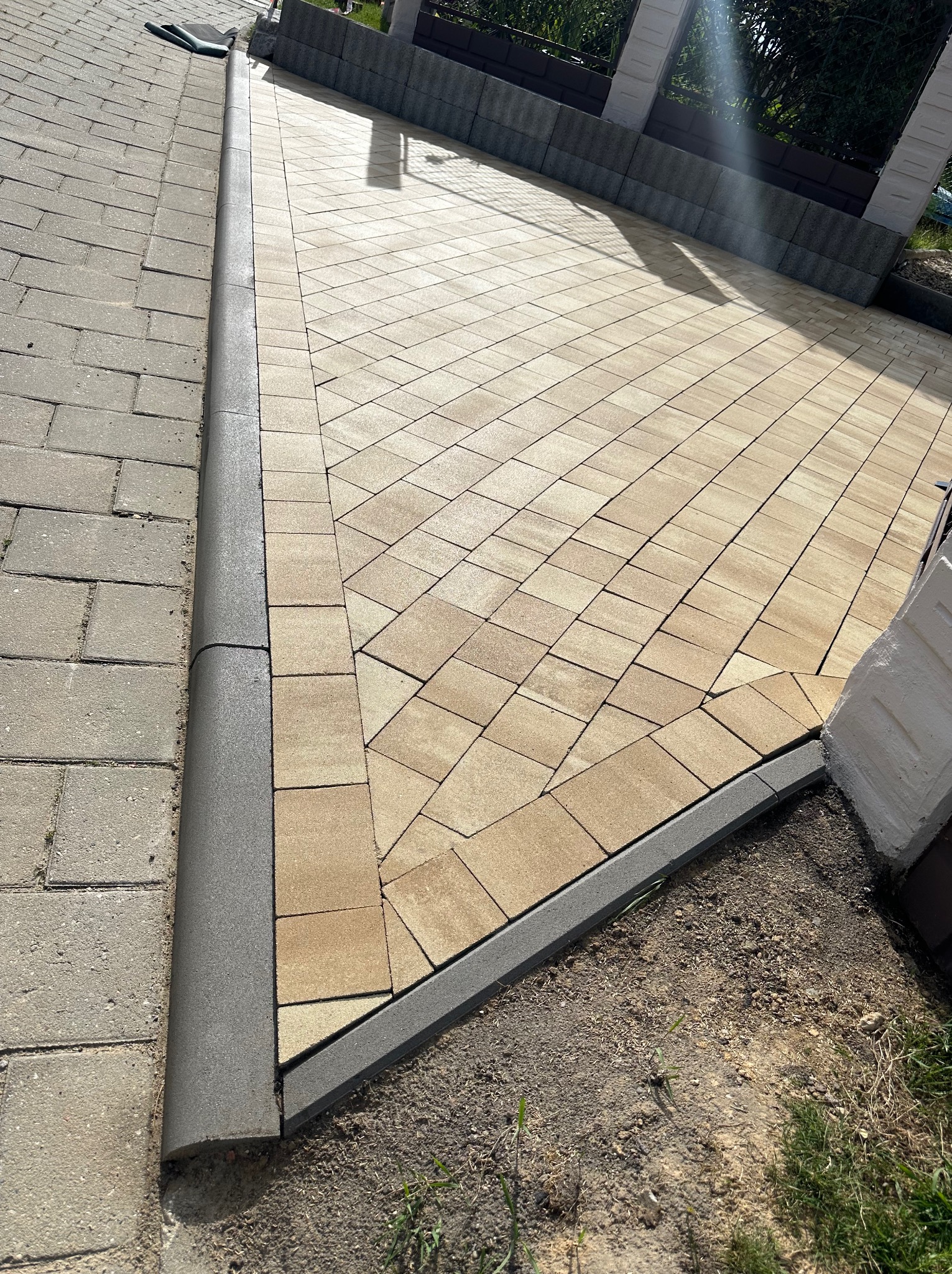 Nowo ułożona, beżowa kostka brukowa z szarą obwódką, płynnie przechodząca w starszą, szarą nawierzchnię; widoczny fragment ogrodzenia w tle.
