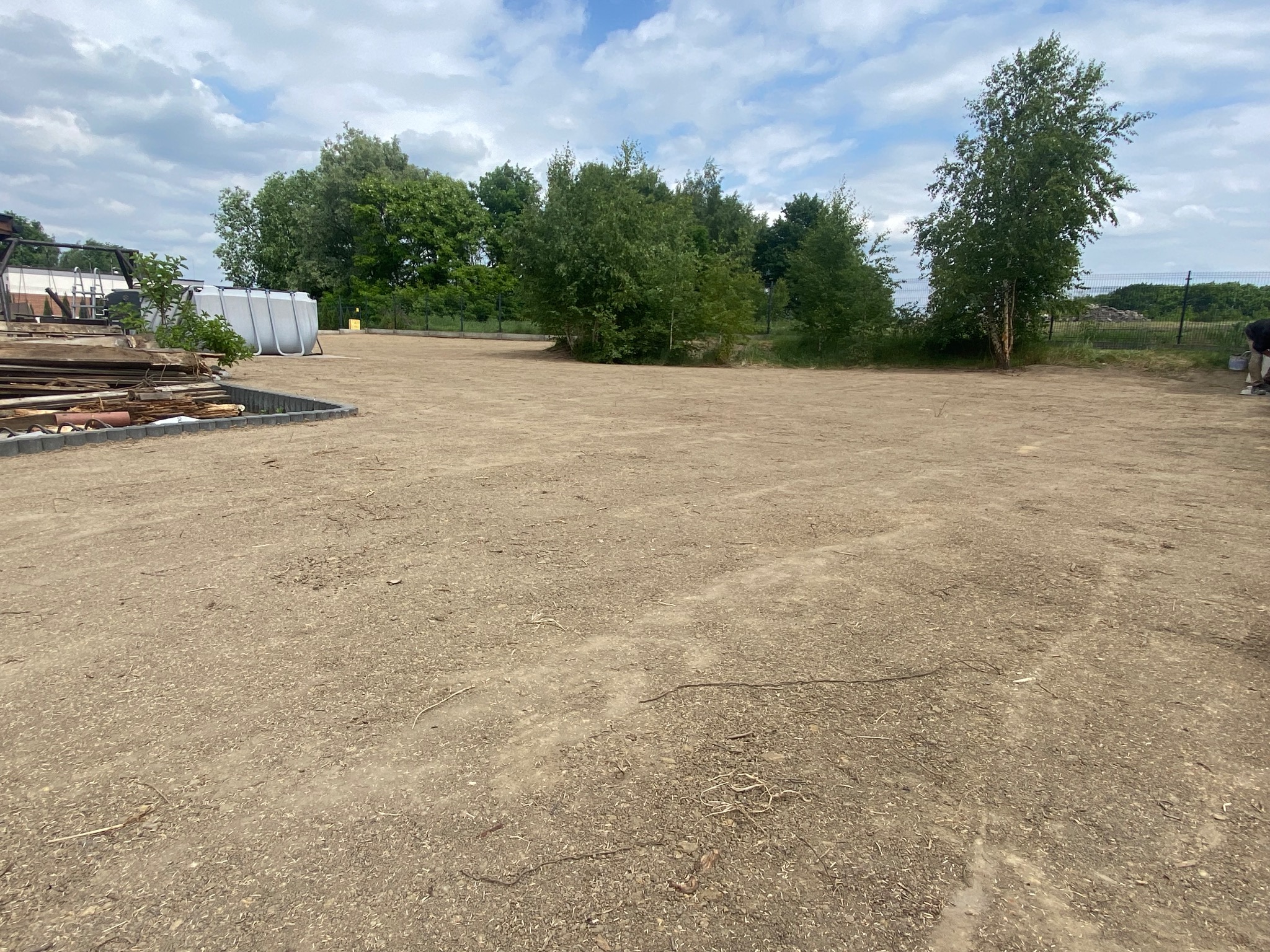 Przygotowany teren pod budowę lub dalsze prace, widoczne obrzeża, drzewa w tle i błękitne niebo z chmurami.