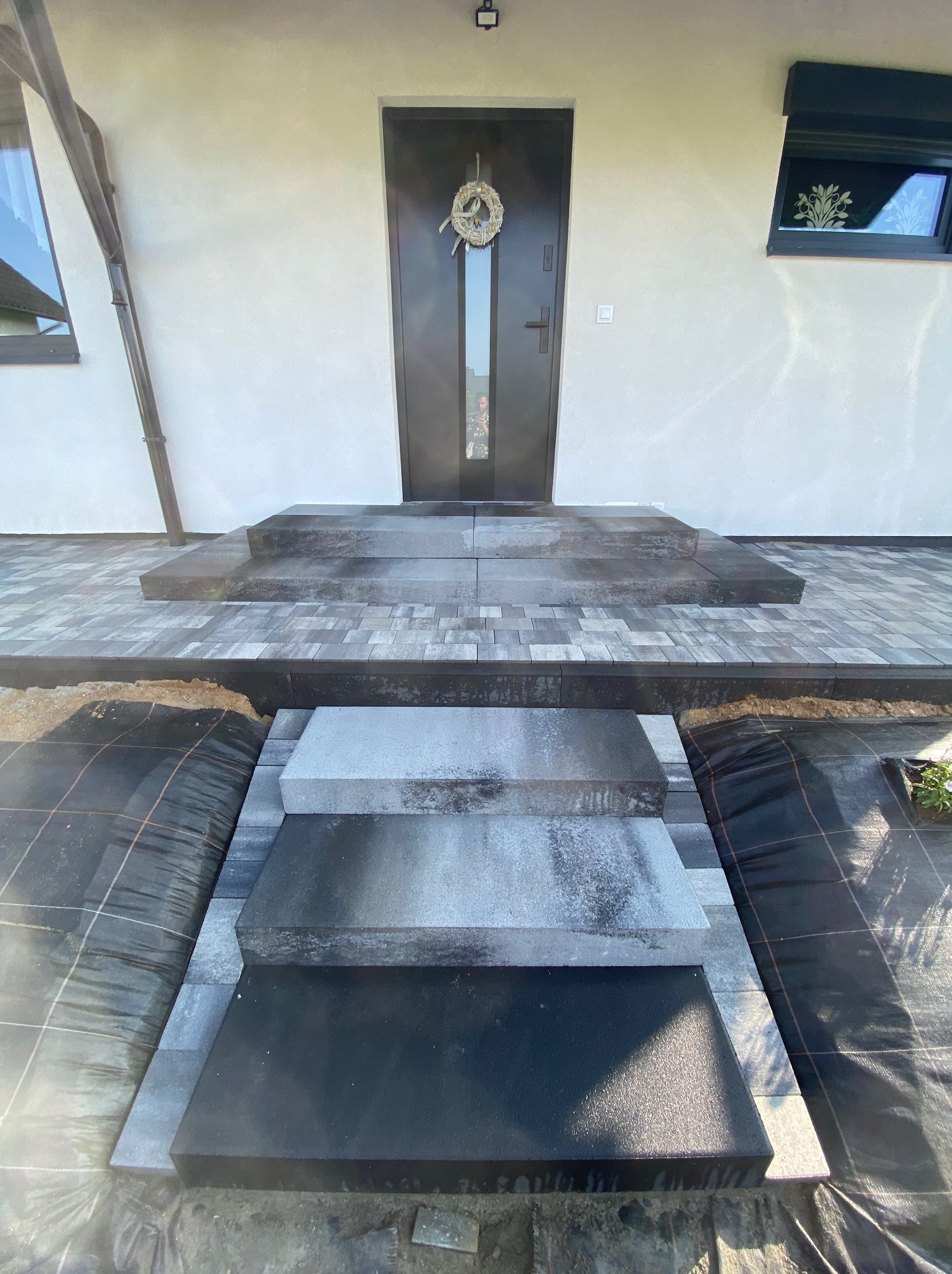 Schody wejściowe z ciemnej kostki brukowej prowadzące do drzwi z wieńcem, widoczne fragmenty włókniny pod kostką.