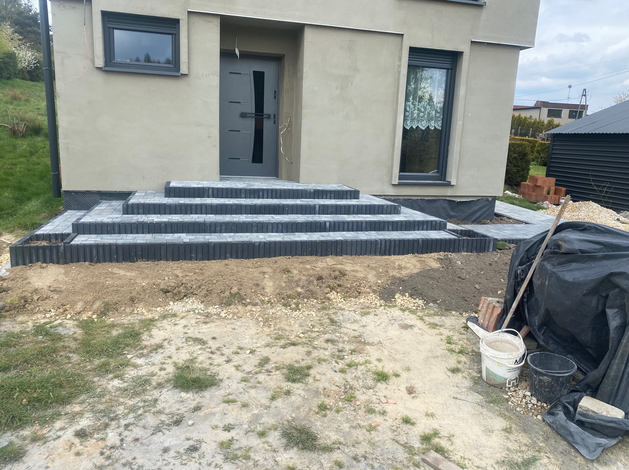 Ułożone schody wejściowe z kostki brukowej w kolorze grafitowym, z widocznymi obrzeżami, prowadzące do nowoczesnego domu z szarą elewacją i stolarką okienną, w tle widoczne są narzędzia i materiały...