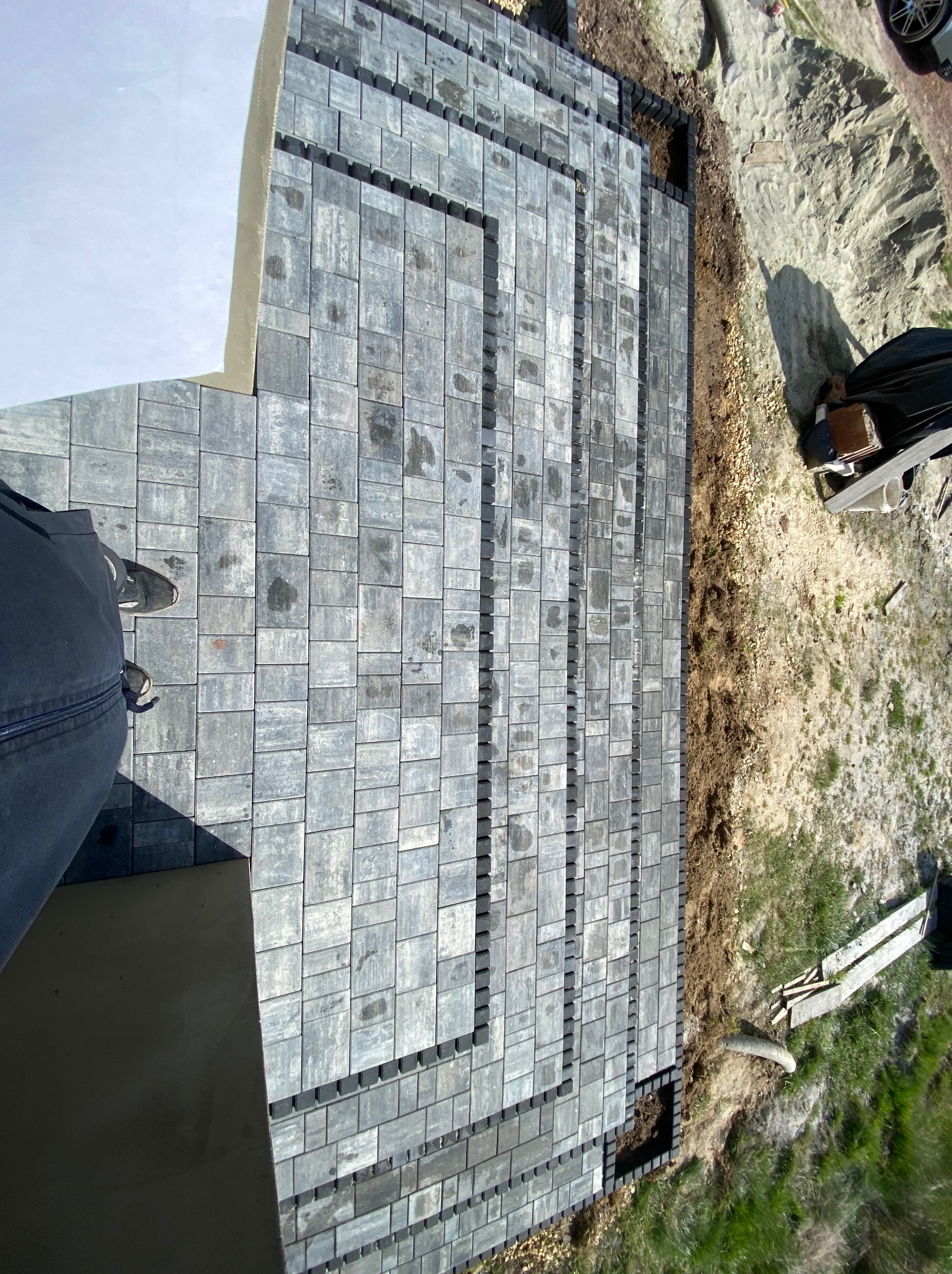 Widok z góry na świeżo ułożoną, szarą kostkę brukową tworzącą schody i chodnik, z widocznymi elementami mocującymi na krawędziach i obrzeżem z ziemi oraz fragmentem buta osoby fotografującej...