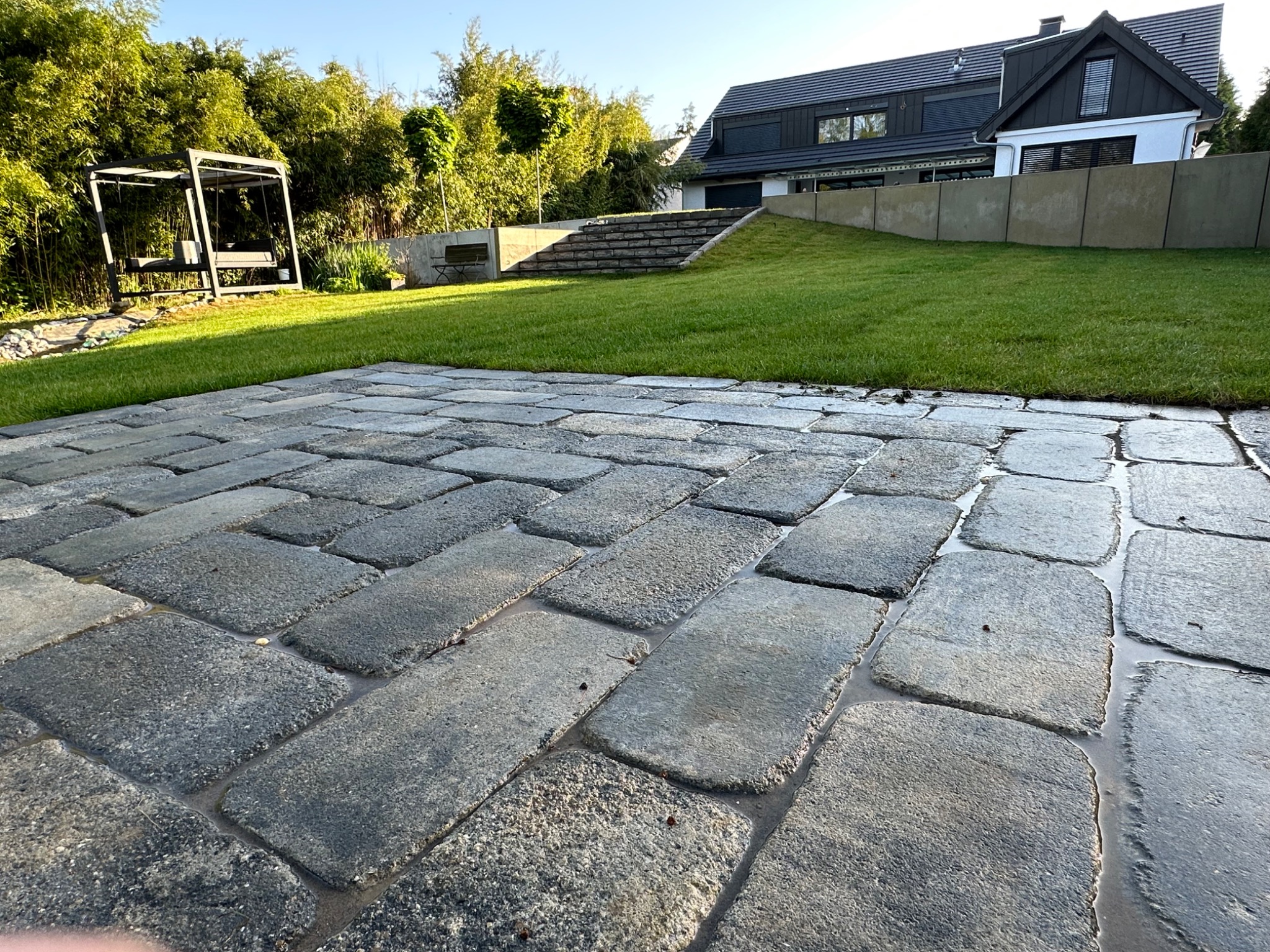 Ułożona szara kostka brukowa na posesji z nowoczesnym domem w tle, widoczne zielone trawniki i elementy architektury ogrodowej, po deszczu, z lekkimi kałużami w spoinach.